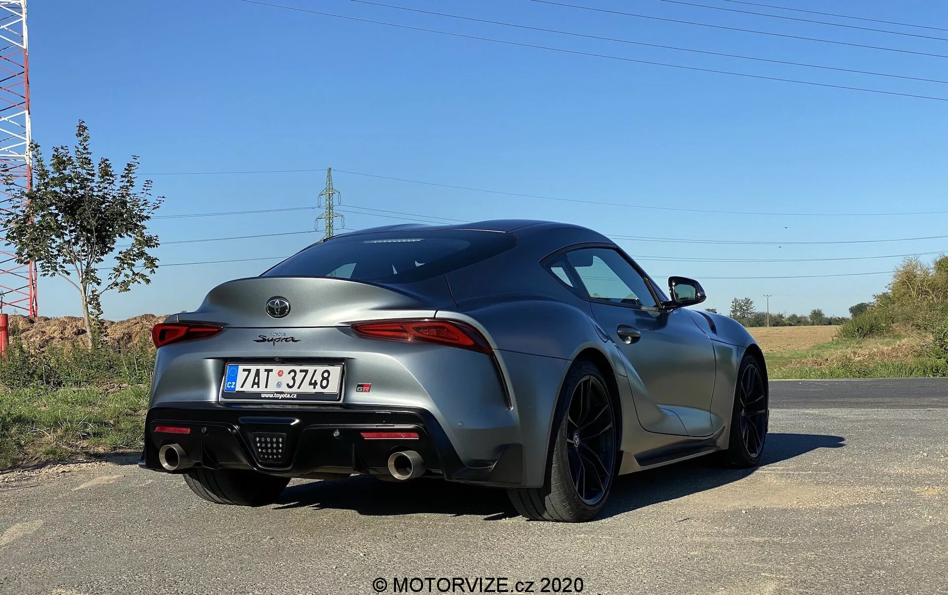 Tylny kąt sportowego samochodu Toyota Supra 2019 2.0 Pure Automatic w kolorze metalicznym szarym, prezentujący agresywny i sportowy design tylnej części, wąskie tylne światła LED skierowane do środka, podwójny układ wydechowy umieszczony centralnie w dyfuzorze, ciemne felgi aluminiowe, europejski styl tablicy rejestracyjnej oraz oznaczenie modelu na bagażniku.