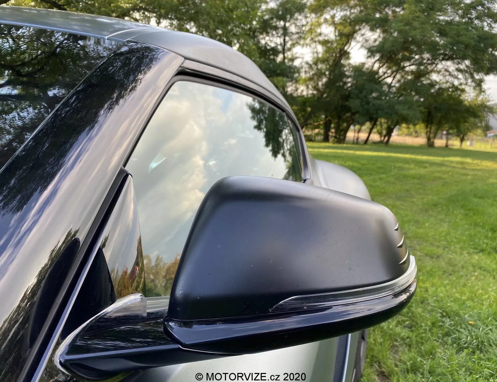 Zbliżenie na eleganckie, czarne lusterko boczne w Toyocie GR Supra 2.0 Pure Automatic z 2019 roku, ukazujące jego nowoczesny, opływowy design. Lusterko osadzone jest w czarnej obudowie o kształcie zoptymalizowanym do zmniejszenia oporu powietrza i jest prawdopodobnie elektrycznie regulowane. Widoczne są przyciemniane boczne okno samochodu i bezszwowy słupek B. W tle znajduje się naturalne otoczenie z trawą i drzewami. Refleksy sugerują jasne warunki na zewnątrz.