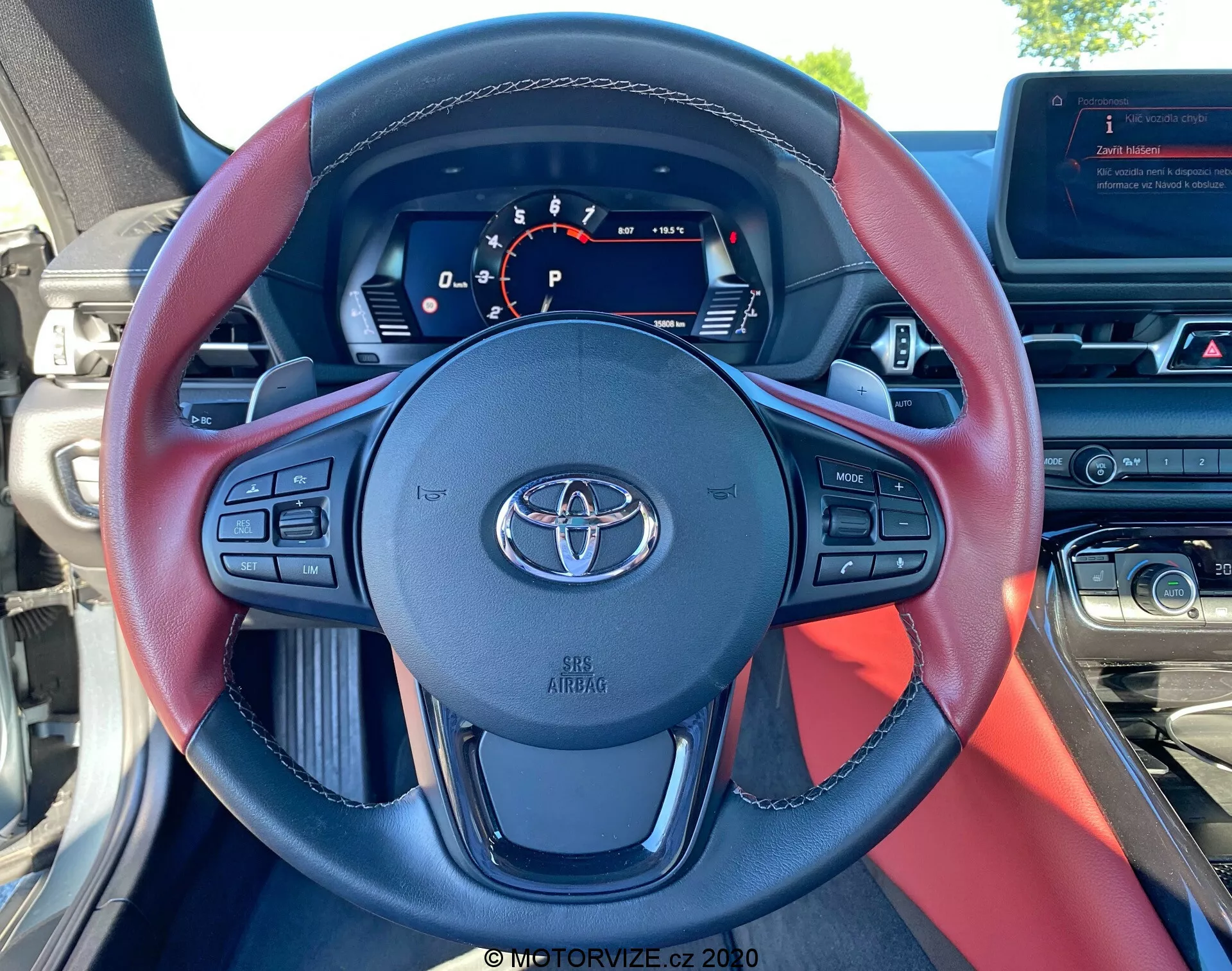 Widok wnętrza z fotela kierowcy Toyoty Supra GR 2019 2.0 Pure Automatic, ukazujący trójramienną kierownicę z czarnymi i czerwonymi akcentami, emblemat Toyoty oraz różne przyciski sterowania do regulacji głośności, komendy głosowe, operacje telefoniczne i adaptacyjny tempomat. Cyfrowy panel instrumentów wyświetla duży centralny prędkościomierz, wskaźnik biegu pokazujący 'P' oraz inne wskaźniki statusu, z widocznym wskaźnikiem paliwa po prawej stronie na górze. Widać również część konsoli środkowej i ekranu infotainment, a także górną część deski rozdzielczej i światło dzienne przez przednią szybę.