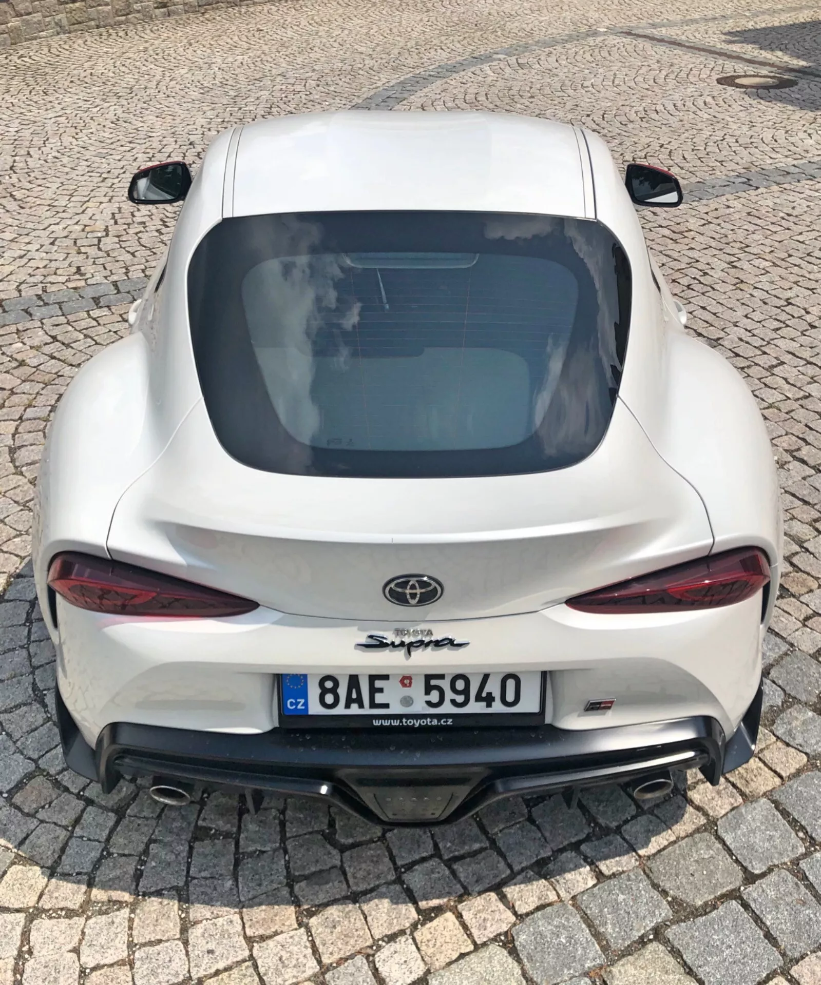 Ten obrazek przedstawia tylny widok białego sportowego samochodu Toyota Supra 2019 z bliska, z wysokiego kąta, pokazując podwójne sporty wydechowe, aerodynamiczny design z zintegrowanym spojlerem, eleganckie tylne światła LED, duży tylny dyfuzor, opadającą linię dachu, wyeksponowane logo Toyota i nazwę modelu 'Supra' oraz tablicę rejestracyjną z Czech. Błyszczące wykończenie lakieru i nowoczesne elementy technologiczne, takie jak światła LED i pakiet aerodynamiczny, wskazują na wydajny i stylowy pojazd w miejskim otoczeniu.