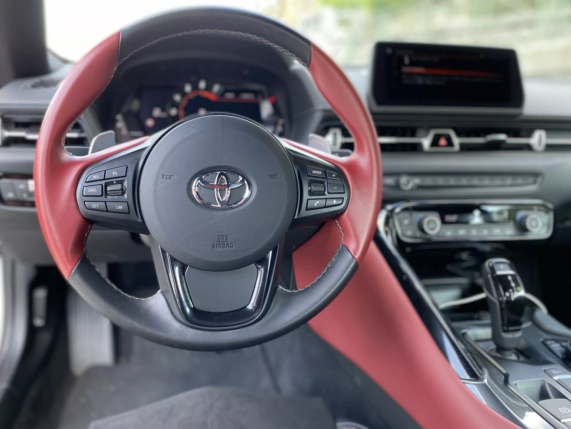 Widok wnętrza Toyota Supra 2.0 Pure Automatic z 2019 roku. Nowoczesna, trzyramienna kierownica z logo Toyota i kolorystyką czerwono-czarną dominuje na obrazie. Zaawansowana kierownica oferuje sterowanie systemem audio, tempomatem i komunikacją bezprzewodową. Za nią widać częściowo cyfrowy zestaw wskaźników, wskazujący na elegancki i technologicznie zaawansowany projekt. Konsola środkowa mieści system infotainment i przyciski sterowania klimatem. Przegląd przez fotel kierowcy z czerwono-czarną skórą i ergonomiczną dźwignią zmiany biegów sugeruje automatyczną skrzynię biegów. Światło dzienne oświetla stojący samochód, podkreślając jego sportowy i luksusowy wygląd wnętrza.
