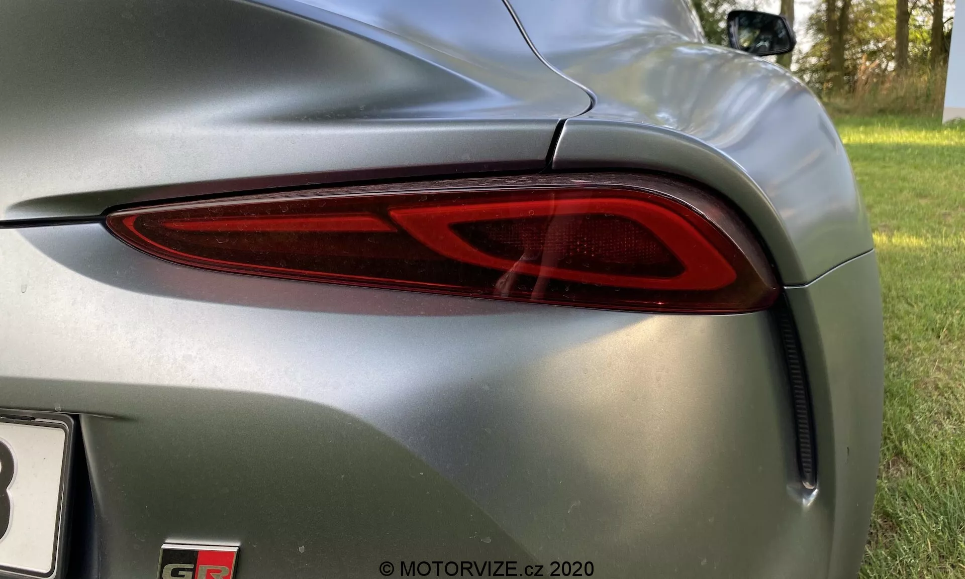 Zbliżenie na tylnią lewą stronę Toyota GR Supra 2019, z nowoczesnym, eleganckim tylnym światłem z technologią LED. Czerwona przezroczysta osłona zakrzywia się wokół boku, zwiększając widoczność. Otaczająca karoseria jest metalicznie szara z lśniącym wykończeniem, odbijającym otoczenie. Tylny zderzak poniżej światła ma otwór wentylacyjny, dodając do aerodynamicznego designu. Widoczna jest część emblematu modelu, sugerując wydajny lub luksusowy pojazd. Zdjęcie zrobione z lekkiego, pochylonego kąta, koncentrując się na elementach designu samochodu.