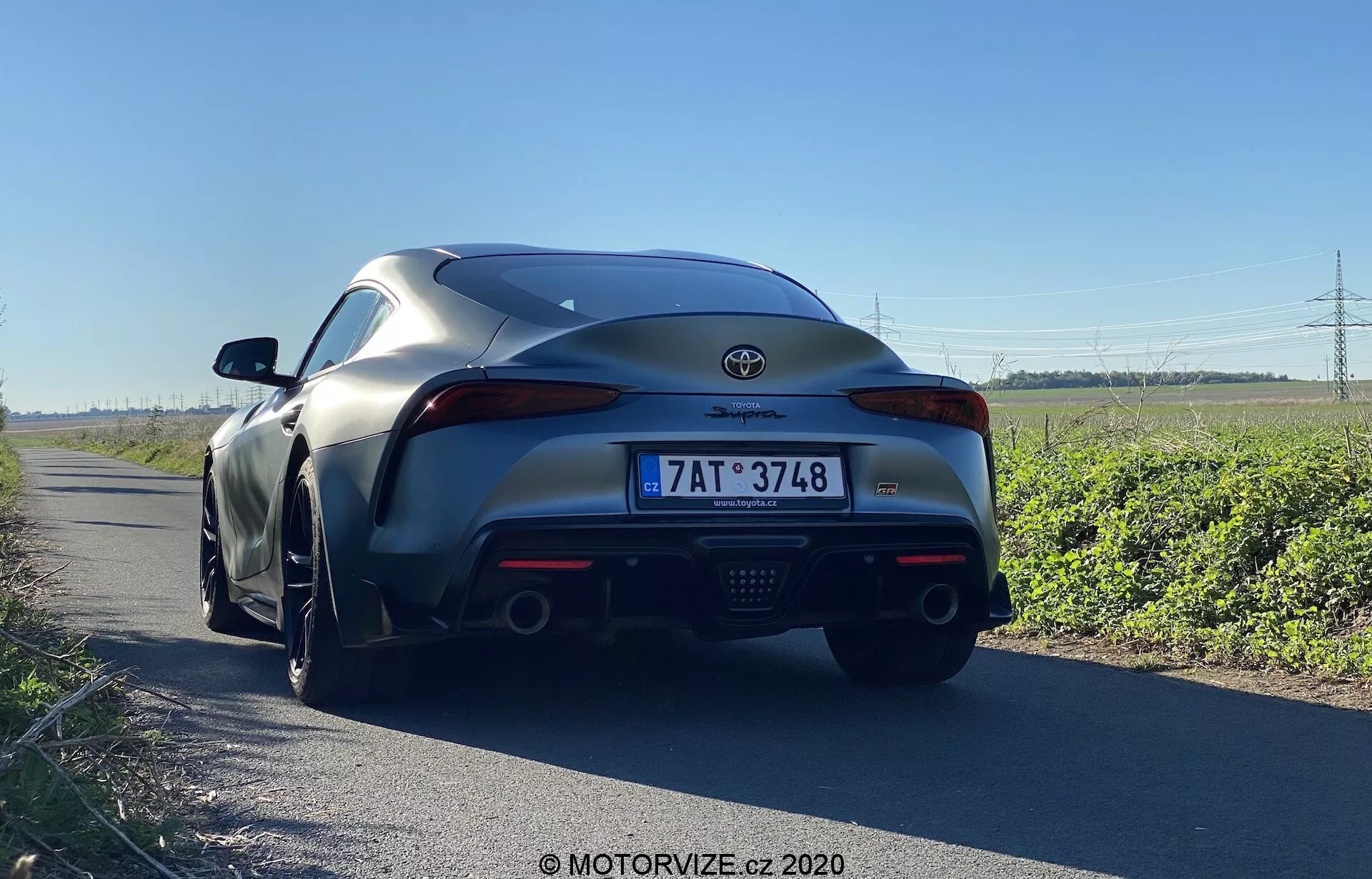 Perspektywa tylna Toyoty Supra 2019, zrobiona z niskiego kąta blisko poziomu gruntu, ukazująca tylny design samochodu. Szczegółowy widok na tylni zderzak, tylne światła, emblemat producenta, podwójne rury wydechowe i tylny dyfuzor. Sceneria na zewnątrz przy drodze, podkreślająca wydajność i nowoczesne elementy wzornictwa, takie jak tylne światła LED, tylny spojler aerodynamiczny i zintegrowany dyfuzor.