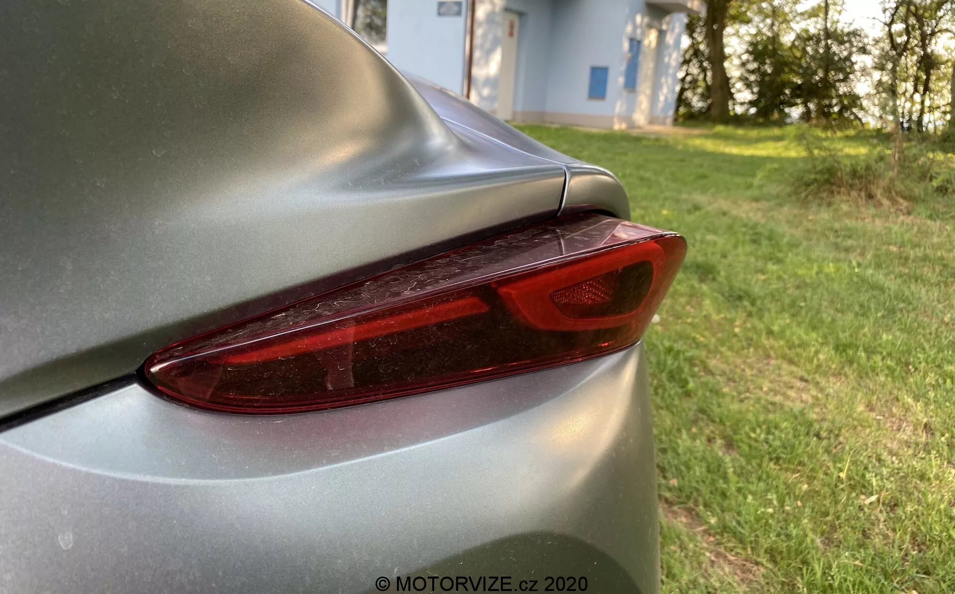 Zbliżenie tylnej sekcji Toyoty Supra 2019 (Toyota-GR-Supra-2-0-Pure-Automatic) prezentującej prawe tylne światło i część pokrywy bagażnika. Nowoczesny design tylnego światła LED charakteryzuje się wyraźnymi, segmentowanymi liniami pod czerwoną osłoną, uzupełnionymi srebrnym lub jasnoszarym nadwoziem z błyszczącym wykończeniem lakieru. Aerodynamiczny kontur i lekko uniesiona część na krawędzi pokrywy bagażnika podkreślają elegancki design samochodu. Zdjęcie koncentruje się na szczegółach tylnego światła na tle bokeh, sugerującym naturalne, spokojne otoczenie na zewnątrz, prawdopodobnie w pobliżu obszaru mieszkalnego.