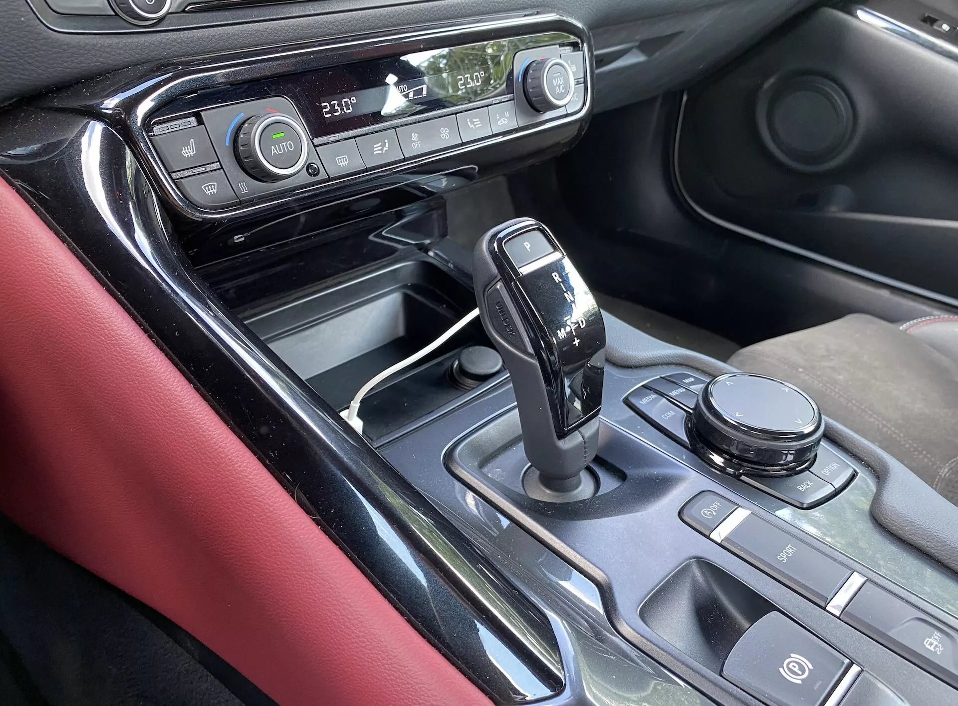 Zbliżony, kątowy widok konsoli centralnej i części deski rozdzielczej Toyota Supra 2019, koncentrujący się na obszarze zmiany biegów. Widok z miejsca pasażera podkreśla nowoczesny dźwignię automatycznej skrzyni biegów z błyszczącym czarnym wykończeniem i chromowanymi detalami w pozycji 'park'. Konsola centralna ma metalowe wykończenie, różne przyciski, selektor trybu jazdy lub pokrętło sterowania systemem infotainment, wyświetlacz pokazujący ustawienie temperatury klimatyzacji, zegar i materiały, takie jak błyszczący czarny, metalowe akcenty i matowe czarne powierzchnie. Czerwone skórzane wykończenie na wewnętrznej stronie drzwi dodaje kontrastu wnętrzu. Obraz sugeruje zaawansowaną technologię infotainment i kontroli klimatu, ergonomiczne rozmieszczenie elementów sterujących i luksusowe elementy wzornictwa.