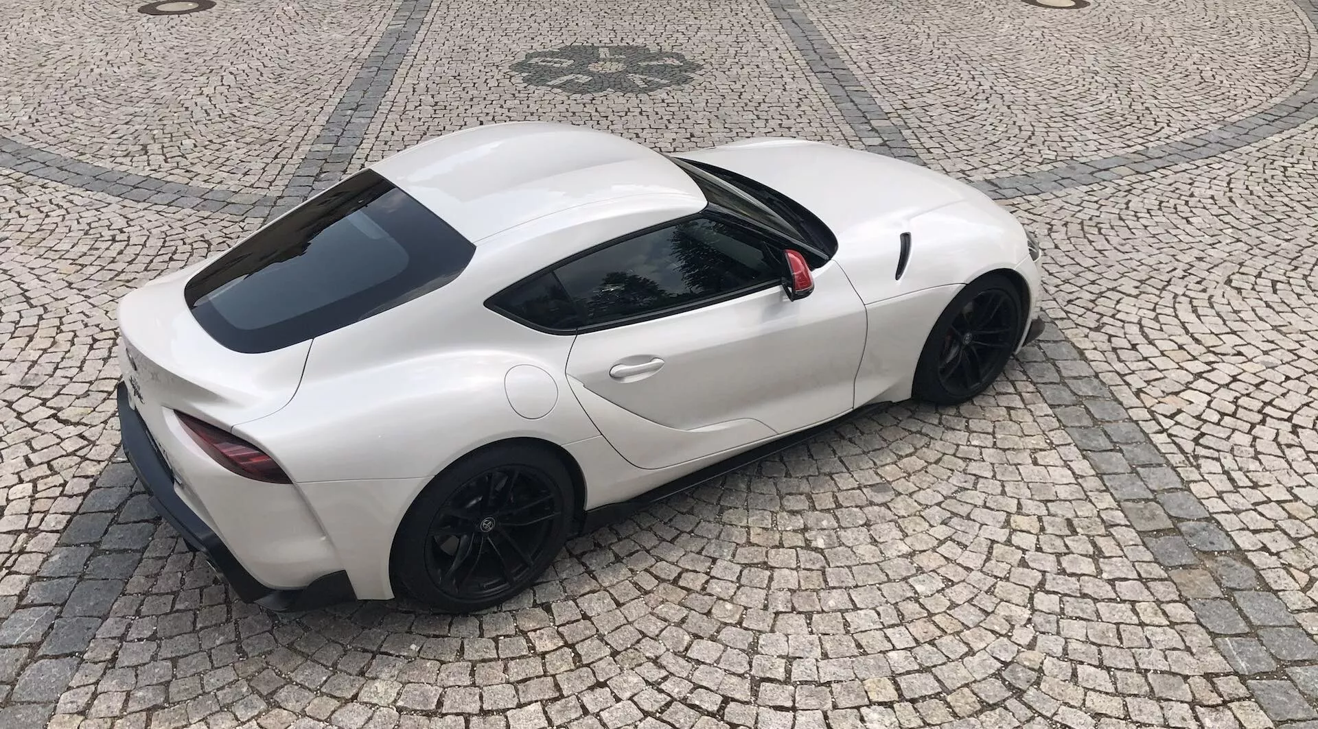 Widok z góry na elegancki, biały samochód sportowy Toyota Supra GR 2.0 Pure Automatic z 2019 roku o aerodynamicznym designie, nadwoziu coupé z dwoma drzwiami i czarnymi felgami aluminiowymi. Czarne lusterka boczne i widoczne kratki wentylacyjne za drzwiami sugerują wysoką wydajność i optymalny rozkład masy. Podwójne końcówki wydechu i mała tylna szyba widoczne, zaparkowany na brukowanej nawierzchni.