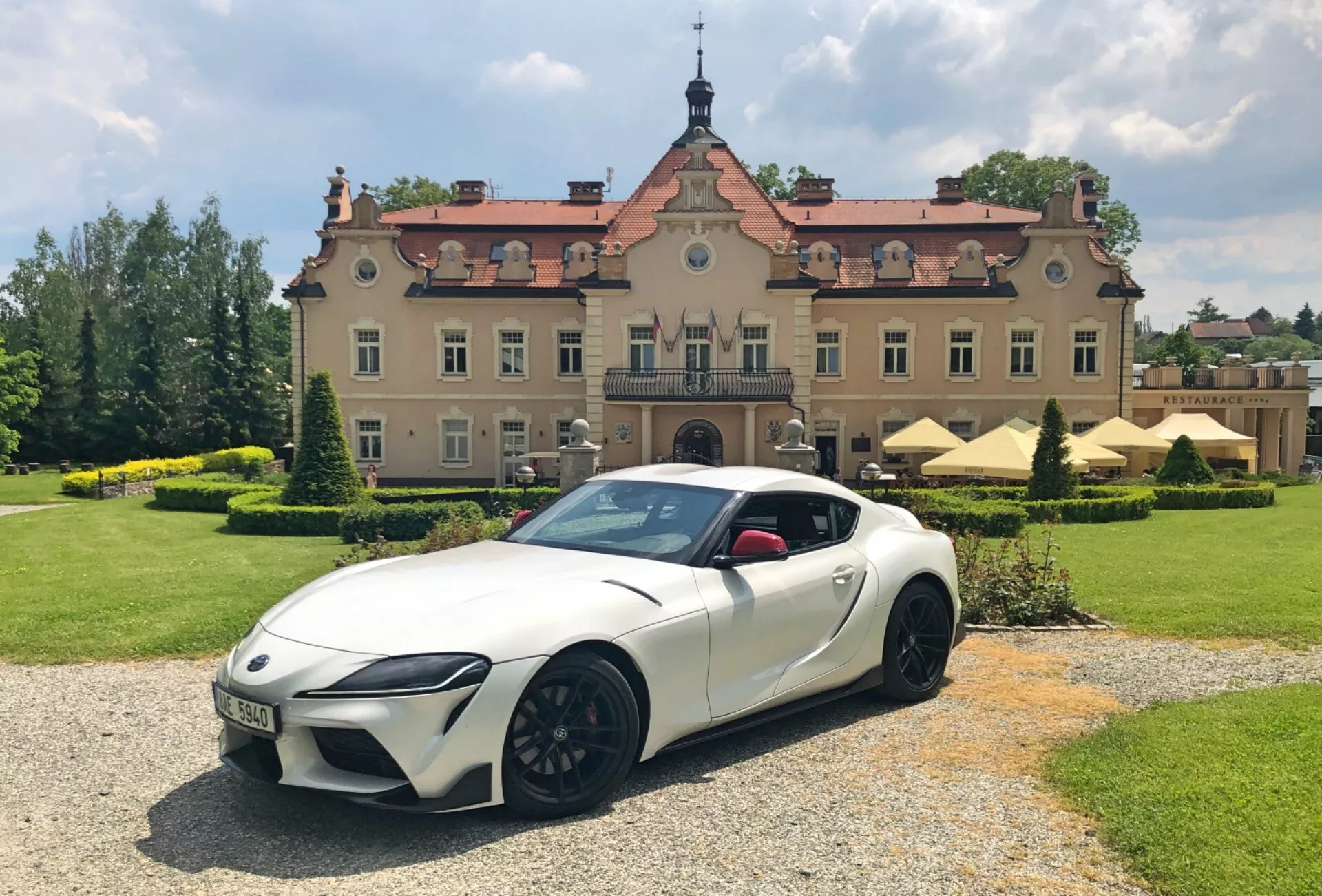 Sportowy samochód Toyota GR Supra 2.0 Pure Automatic z 2019 roku w perłowej bieli z czarnymi akcentami i wieloramiennymi, aluminiowymi felgami, zaparkowany na żwirowej powierzchni z okazałym budynkiem w tle. Obraz uchwycony z niskiego, przedniego kąta trzy-czwartego, pokazuje ostre reflektory LED, agresywny styl zderzaka, opadającą linię dachu coupe i wyraźne nadkola. Otoczenie obejmuje zadbane tereny zielone pod częściowo zachmurzonym niebem, co dodaje luksusowego i dynamicznego uroku scenerii.