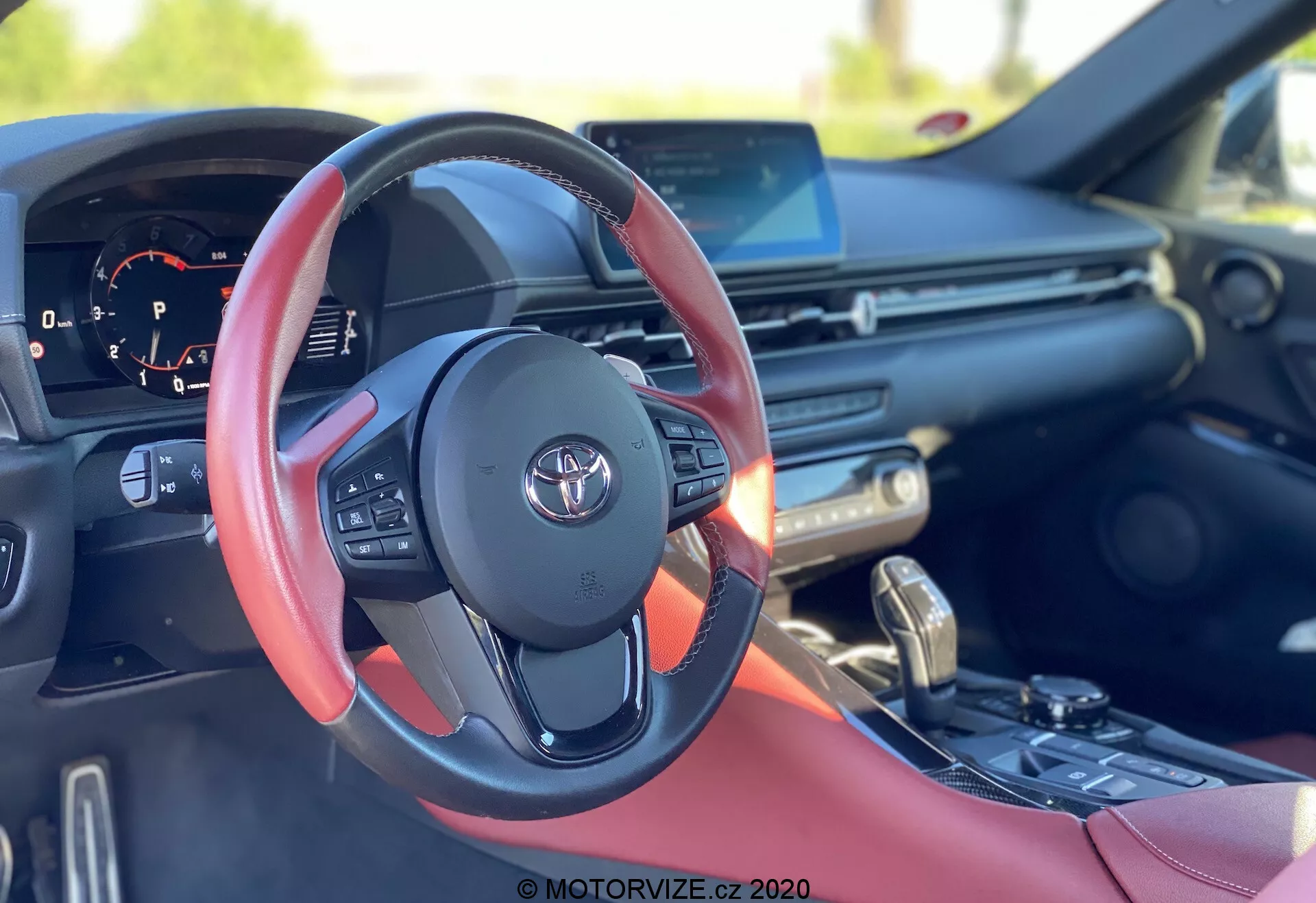 Obraz jest przedstawieniem wnętrza Toyoty Supra 2019 (Toyota-GR-Supra-2-0-Pure-Automatic), skoncentrowanym głównie na skórzanej kierownicy z czerwonymi akcentami, z widocznym logiem Toyota. Perspektywa z siedzenia kierowcy pokazuje różne kontrolki, w tym przyciski do sterowania dźwiękiem, telefonem bez użycia rąk i ustawieniami tempomatu. Łopatki za kierownicą sugerują obecność półautomatycznej skrzyni biegów. Zegar pokazuje obrotomierz i częściowo widoczny, cyfrowy wyświetlacz. Deska rozdzielcza wyposażona jest w szeroki wyświetlacz multimedialny prawdopodobnie obsługujący GPS, odtwarzanie multimediów i integrację smartfona, z nawiewami powietrza i przyciskami do sterowania klimatyzacją poniżej. Konsola środkowa zawiera dźwignię zmiany biegów do automatycznej skrzyni biegów oraz selektor trybu jazdy do regulacji dynamiki jazdy. Wnętrze samochodu ma czerwone skórzane wstawki, co sugeruje sportowy lub luksusowy pakiet.