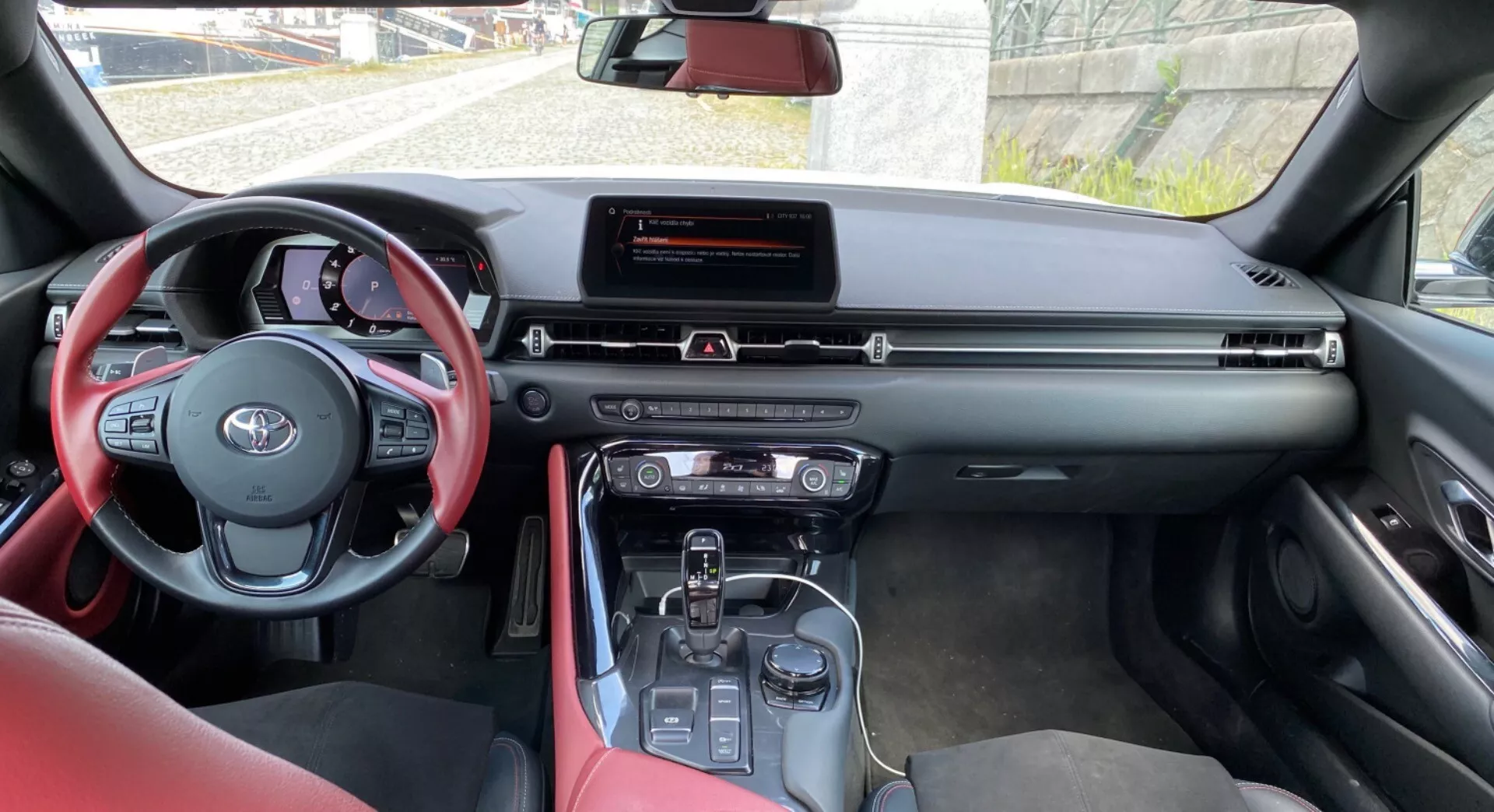Zdjęcie przedstawia wnętrze nowoczesnego pojazdu Toyota, konkretnie Toyota Supra 2019 (Toyota GR Supra 2.0 Pure Automatic), z perspektywy osoby siedzącej na tylnym siedzeniu pasażera, lekko przesuniętej w stronę kierowcy. Na pierwszym planie widnieje kierownica owinięta czerwoną skórą z dotykowymi elementami sterującymi. Centralna deska rozdzielcza posiada panoramiczny wyświetlacz infotainment umieszczony u góry, otoczony dotykowymi przyciskami i potencjalnym wyświetlaczem klimatyzacji. W konsoli środkowej znajduje się selektor zmiany biegów wskazujący na automatyczną skrzynię biegów, co może sugerować technologię hybrydową lub elektryczną. Czerwone krawędzie przednich siedzeń sugerują czerwoną tapicerkę jako cechę estetyczną tej wersji modelu. Dziennie światło oświetla wnętrze samochodu, a widok przez przednią szybę ukazuje budynek i niebo, co potwierdza, że samochód jest zaparkowany.