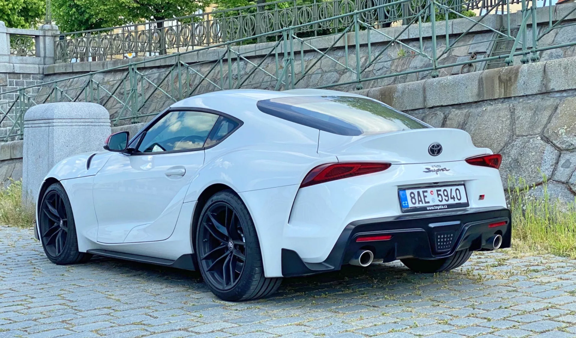 Tylny widok w trzech czwartych białego 2019 Toyota GR Supra 2.0 Pure Automatic, ukazujący podwójny układ wydechowy, zintegrowany tylny spojler, tylne światła LED, sportowe wieloszprychowe felgi, tylny dyfuzor i boczne wloty powietrza. Pojazd jest zaparkowany w miejskim otoczeniu z kamiennym murem i zielenią w tle.