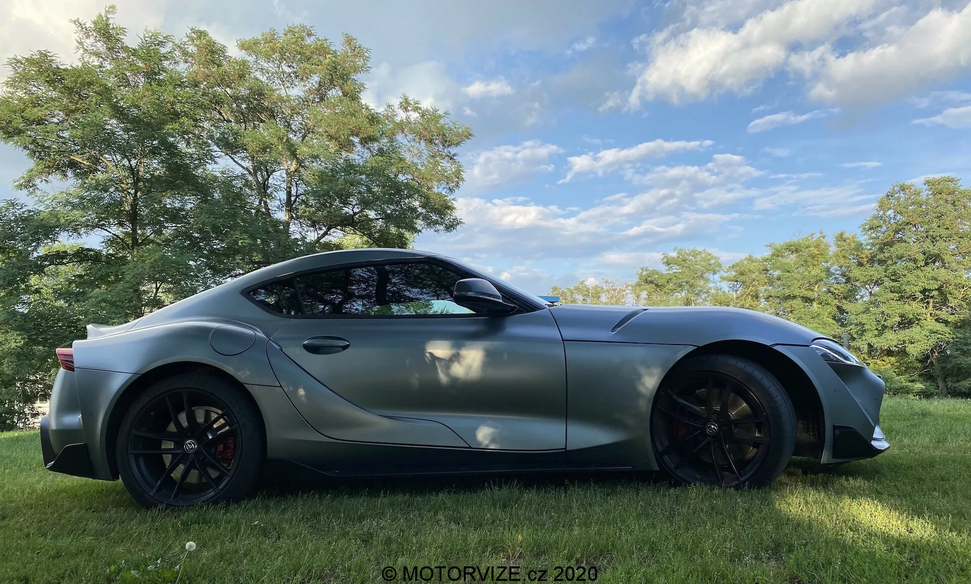 Obraz sportowego samochodu Toyota GR Supra 2.0 Pure Automatic z 2019 roku, uchwyconego z profilu bocznego, ustawionego na zewnątrz na trawiastym polu z drzewami w tle, pod częściowo pochmurnym niebem. Elegancki, aerodynamiczny design obejmuje długą maskę i pochylony tył typu fastback, metaliczne wykończenie, duże felgi aluminiowe z widocznymi zaciskami hamulcowymi, aerodynamiczne lusterka boczne i refleksyjne przyciemniane szyby. Rozszerzone nadkola i linie konturowe podkreślają wydajnościowy charakter nowoczesnego samochodu sportowego.
