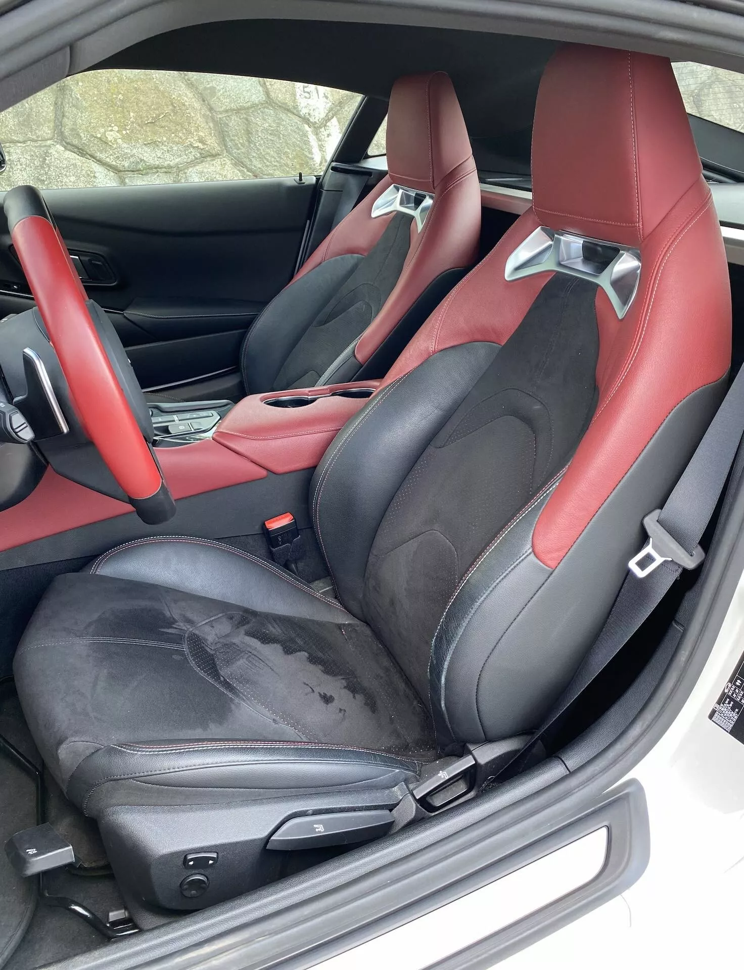 Zbliżenie wnętrza po stronie kierowcy Toyota GR Supra 2.0 Pure Automatic 2019, z sportowymi siedzeniami kubełkowymi w efektownej czarno-czerwonej tapicerce ze skóry. Siedzenia mają zintegrowane zagłówki i boczne wzmocnienia zapewniające wsparcie, widoczne zużycie na krawędzi siedzenia kierowcy oraz elektrycznie regulowane ustawienia siedzeń wskazujące na luksus. Widoczna jest również część wnętrza drzwi i konsoli środkowej z dopasowanym schematem kolorów, a także niestandardowa mata podłogowa i pasy bezpieczeństwa wychodzące z otworów w ramionach.