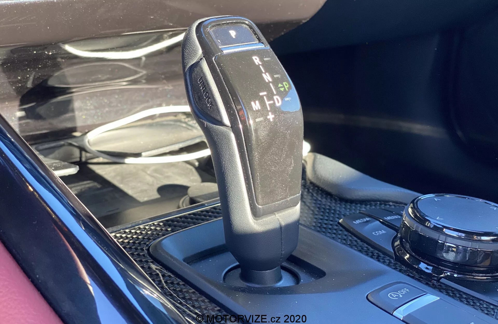 Zbliżenie zdjęcia dźwigni zmiany biegów automatycznej w Toyota GR Supra 2.0 Pure 2019 z opcjami parkowania (P), cofania (R), neutral (N), jazdy (D) i manualnym/sportowym (M), z teksturowanym uchwytem ze skóry i czarnego plastiku na nowoczesnej konsoli środkowej z wykończeniem przypominającym włókno węglowe i pokrętłem do kontroli multimediów.