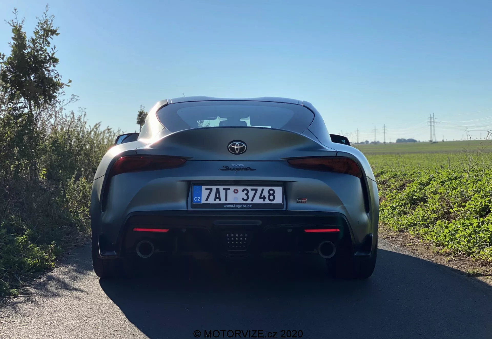 Obraz ten przedstawia tylno-środkowy widok na Toyota GR Supra 2.0 Pure Automatic z 2019 roku, podkreślając kluczowe detale, takie jak emblemat Toyota, oznaczenie Supra, zintegrowany tylny spojler, centralne światło hamowania, eleganckie zespoły tylnych świateł, podwójne końcówki wydechu, europejską tablicę rejestracyjną, błyszczące metaliczne wykończenie lakieru oraz czarną dolną część tylnego zderzaka z elementami przypominającymi dyfuzor.