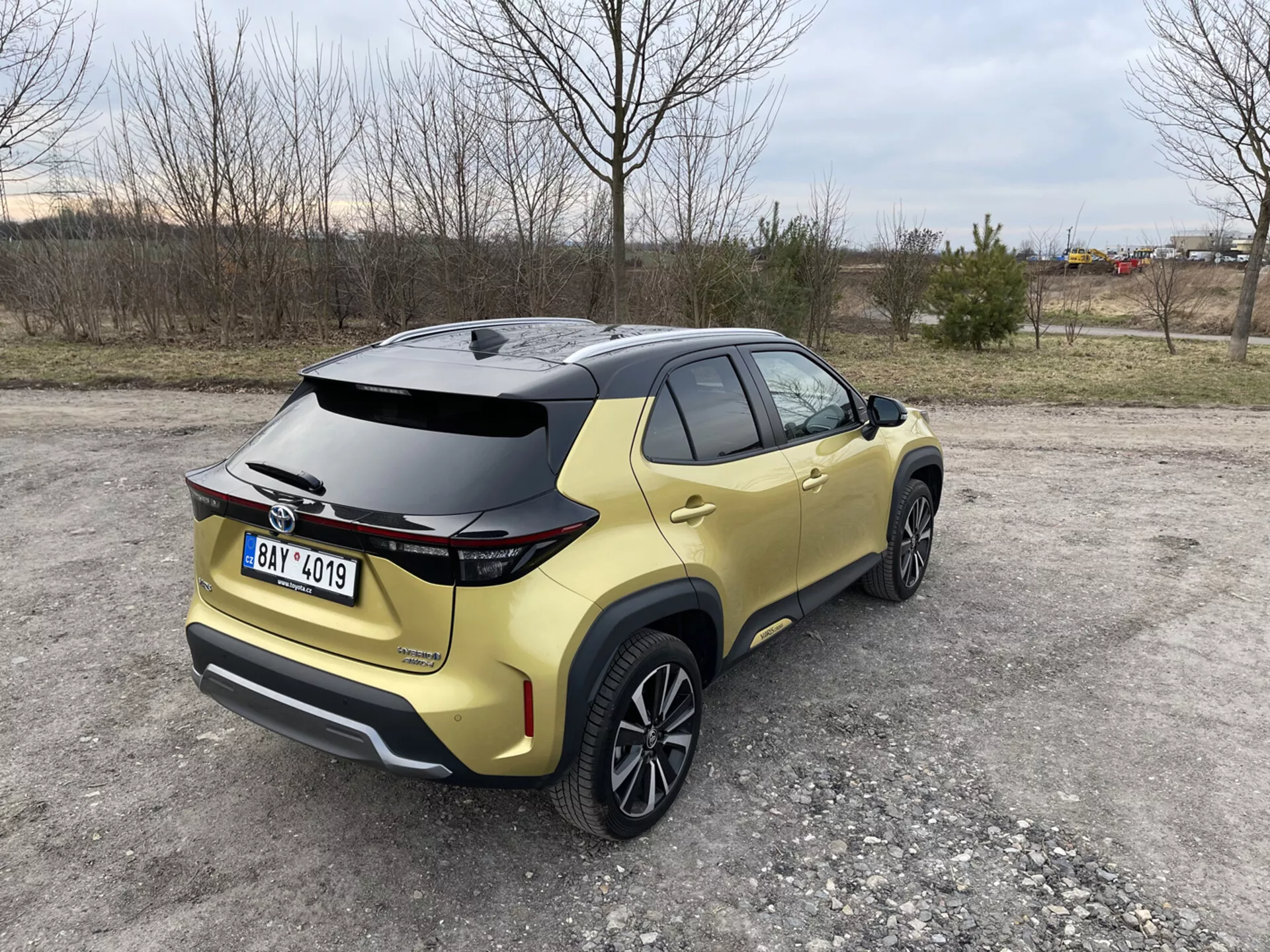 Toyota Yaris Cross 2021 uchwycona z tylnego trójkątnego widoku podkreślającego jej aerodynamiczny spoiler, metaliczne złote malowanie i tylne światła LED, zaparkowana na nieutwardzonej powierzchni z naturalnym i przemysłowym tłem.
