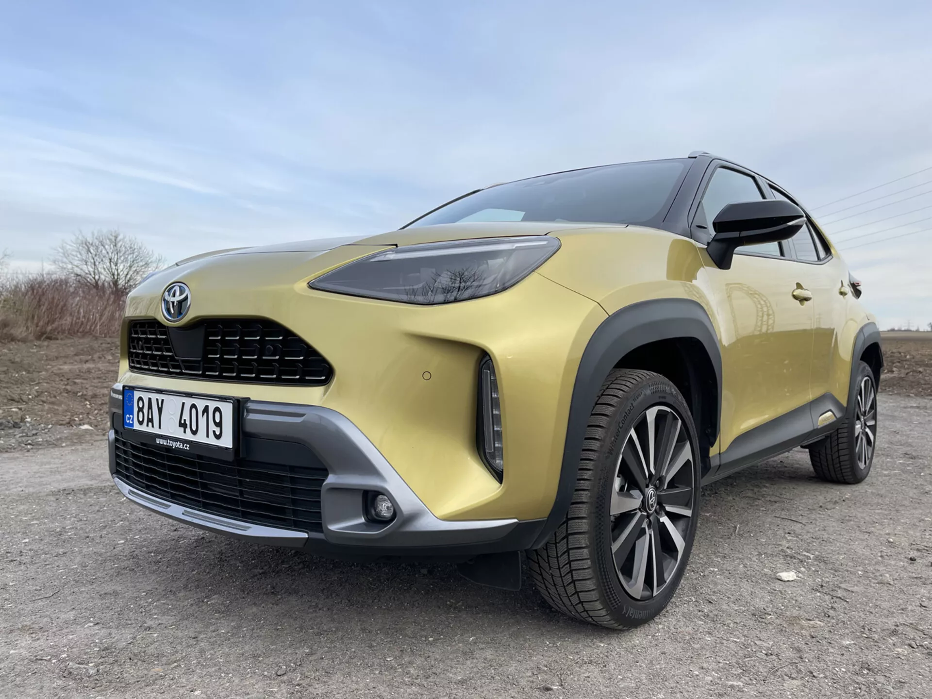 SUV Toyota Yaris-Cross 2021 w kolorze złotym prezentujący swój przedni lewy kwadrant z niskiej perspektywy szerokokątnej podkreślający reflektory LED, grill, zderzak, obręcz ze stopu oraz nowoczesne elementy designu na nieutwardzonej powierzchni