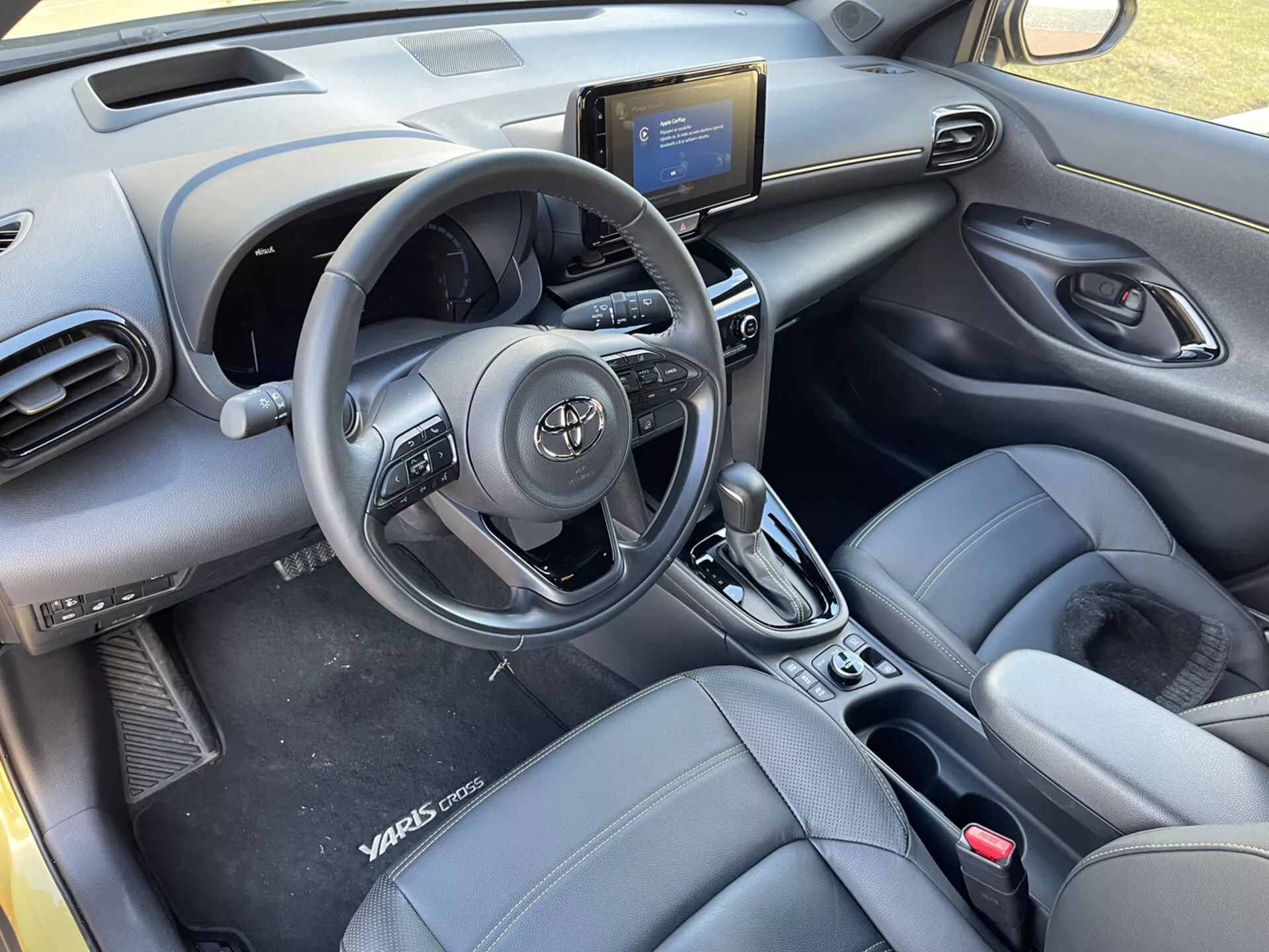 Widok wnętrza Toyoty Yaris-Cross 2021 z tylnych siedzeń, podkreślający kierownicę z przyciskami sterującymi, nawiewy na desce rozdzielczej, konsolę środkową z systemem rozrywki i informacji, kontroli klimatyzacji, dźwignię automatycznej skrzyni biegów, przednie fotele kubełkowe z wyglądem skóry z oznaczeniem Yaris Cross, panele drzwi z kontrolami elektrycznymi, oraz nowoczesny, elegancki wygląd kabiny.