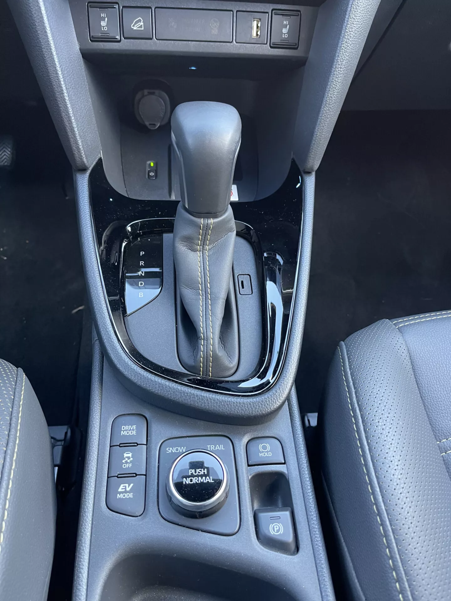 Wnętrze centralnej konsoli Toyota Yaris Cross 2021 z automatyczną dźwignią zmiany biegów w pozycji parkowania, otaczającymi przyciski kontroli dla asystenta parkowania, funkcji utrzymywania hamulca, czujników parkowania i trybów jazdy w tym SPORT, ECO, EV, AUTO i NORMAL, prezentująca materiał przypominający skórę z detalami szwów i błyszczące czarne detale