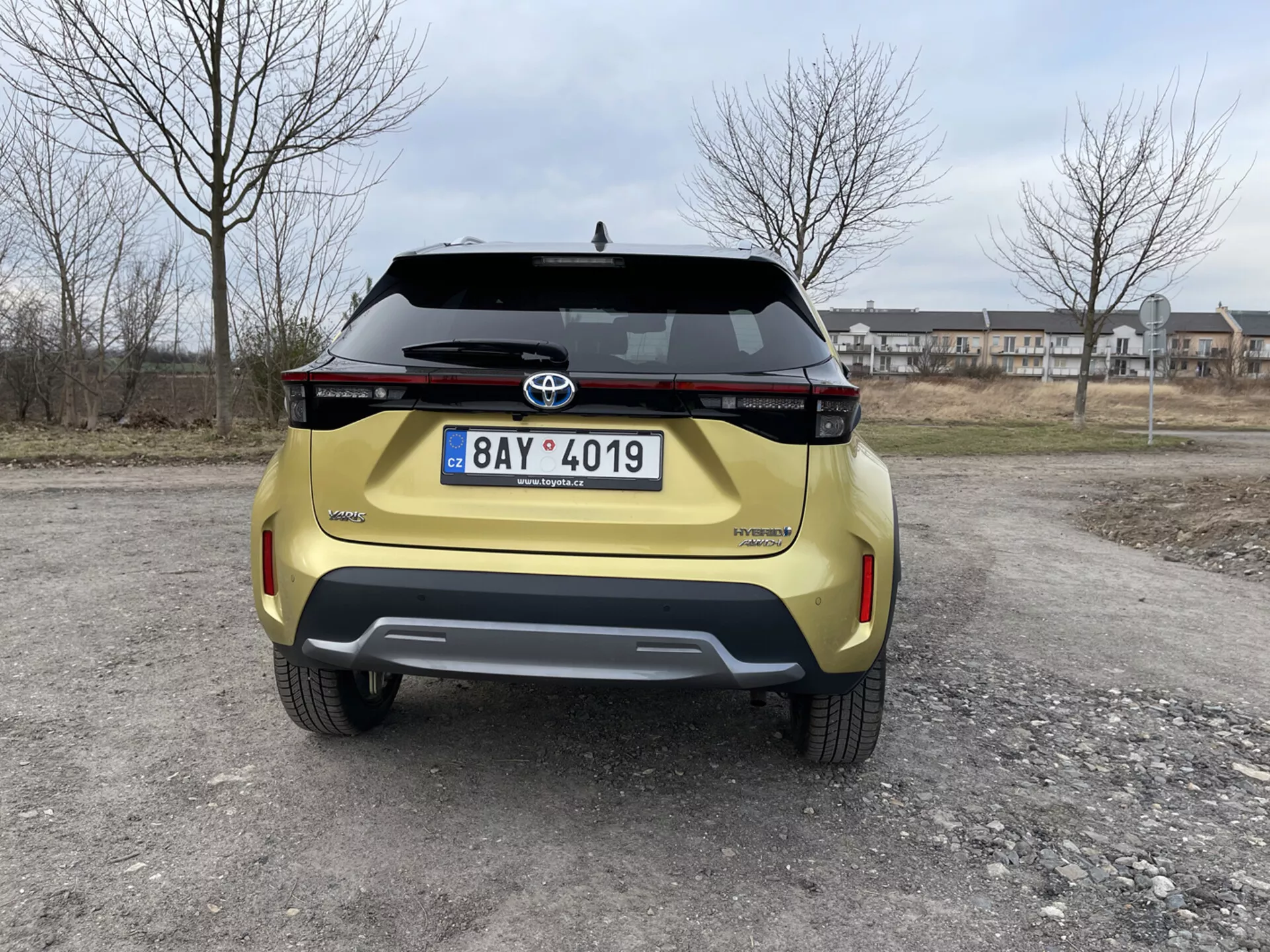 Tylny widok żółtej Toyoty Yaris Cross 2021 uchwycony na wysokości oczu, przedstawiający jej nowoczesny design, poziome zespoły świetlne tylne, emblemat Toyota, nazwę modelu Urban Cruiser, tablicę rejestracyjną, tylny zderzak z elementem kontrastowym z wbudowanymi czujnikami parkowania i klamkę klapy na tle przedmieścia.