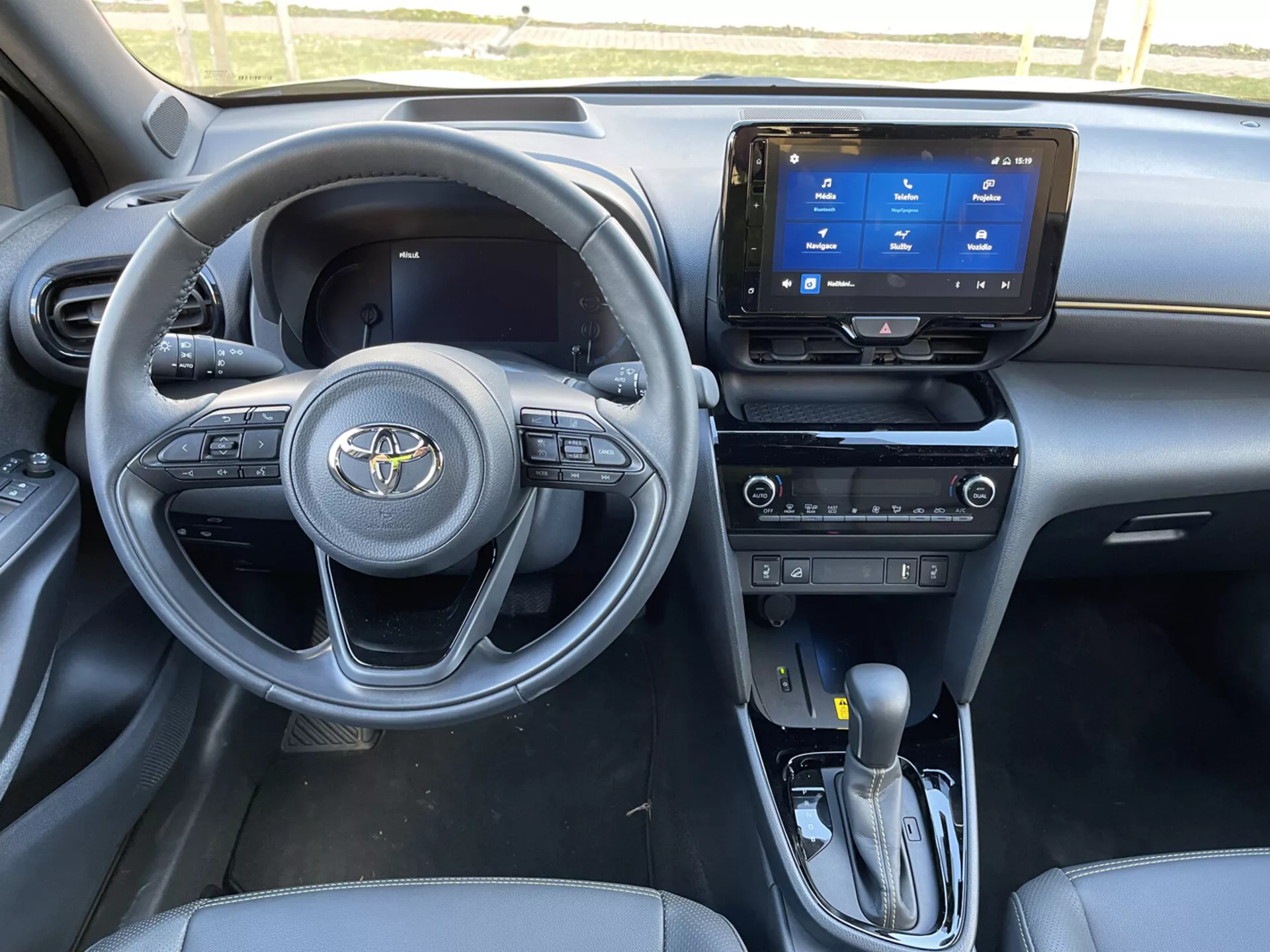 Wnętrze Toyota Yaris Cross 2021 z perspektywy pasażera przedniego, pokazujące deskę rozdzielczą, kierownicę z zamontowanymi sterowaniami, system infotainmentu dotykowego, automatyczną dźwignię zmiany biegów oraz eleganckie cechy siedzeń i projektu w ciemnej kolorystyce