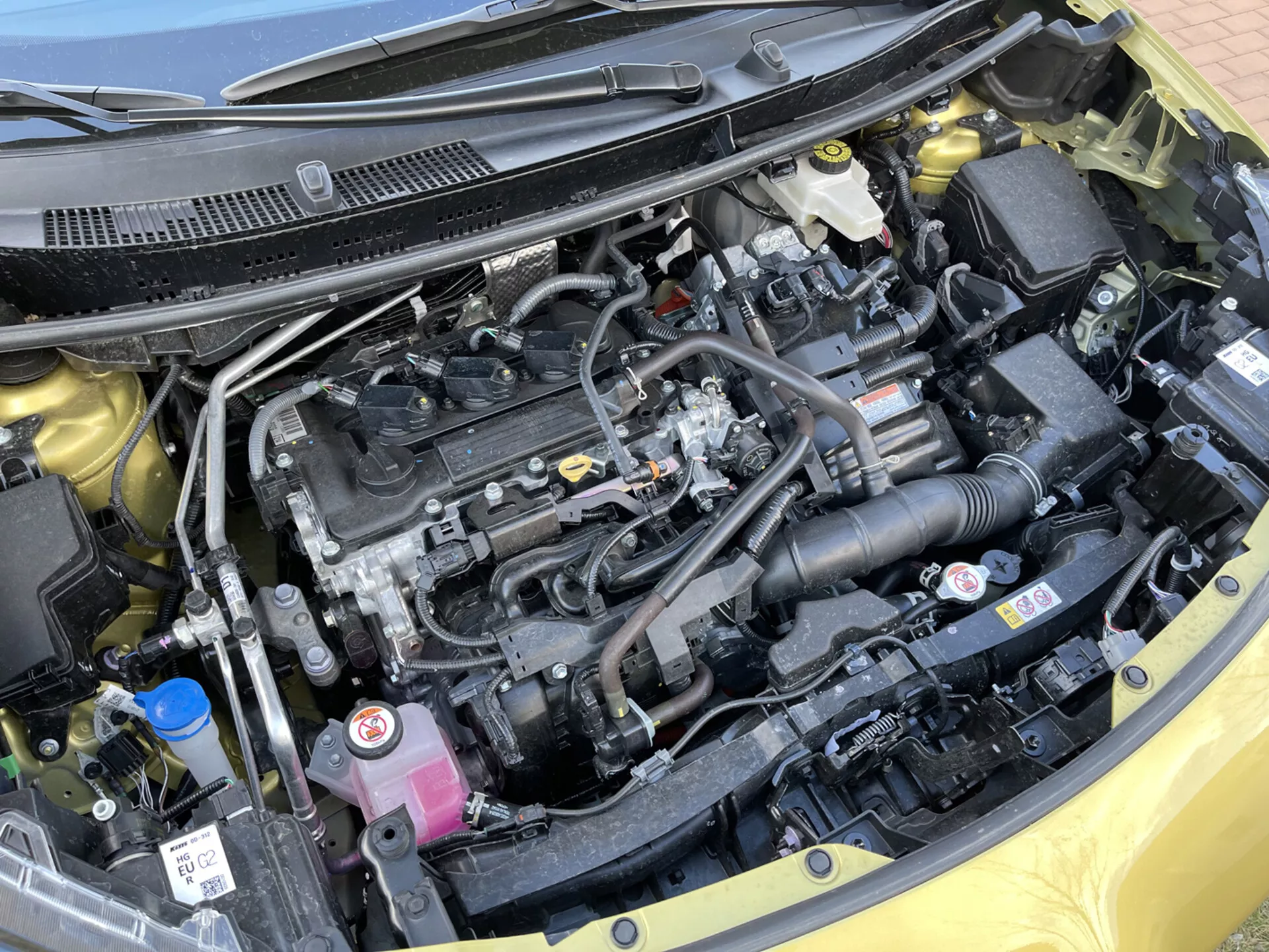 Zbliżony widok komory silnika Toyoty Yaris-Cross 2021 pokazujący silnik rzędowy, czarną plastikową osłonę silnika, system dolotu powietrza, zbiorniki na płyn chłodzący i płyn hamulcowy, komponenty elektryczne, baterię, chłodnicę i starannie zorganizowany układ z etykietami bezpieczeństwa.