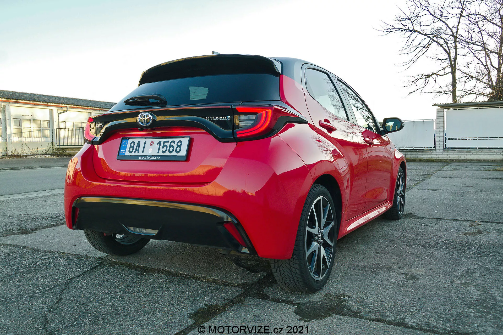 Żywo czerwoną Toyotę Yaris z 2020 roku sfotografowano pod kątem tylno-trzecioczwartym, podkreślającym diodowe światła tylne, tylny spojler, emblemat modelu hybrydowego, wieloramienne obręcze ze stopu metali lekkich oraz widoczne funkcje wspomagania kierowcy, takie jak czujniki parkowania i kamera cofania, uchwycone z perspektywy nieco poniżej wysokości auta.