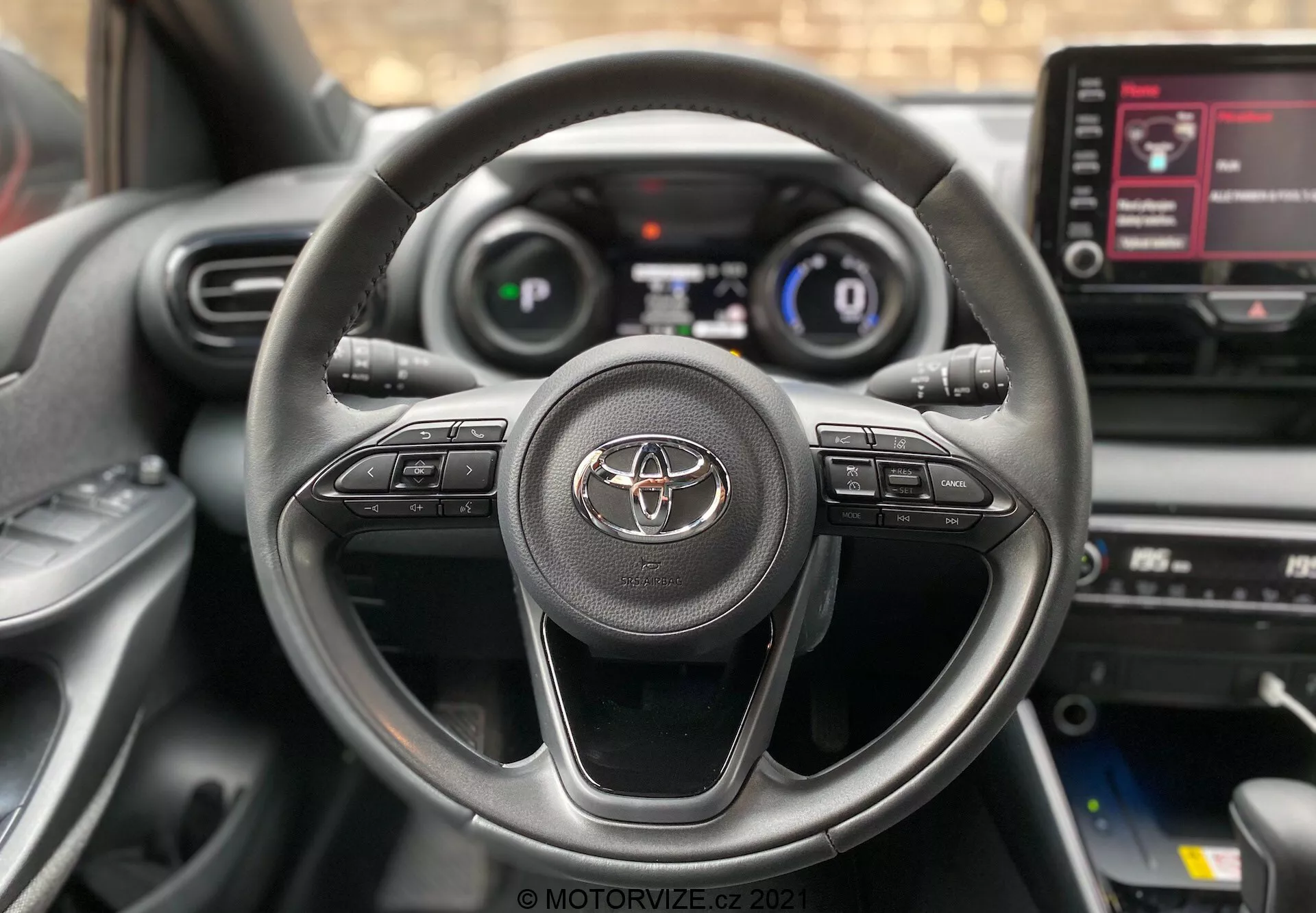 Zbliżenie wnętrza Toyota Yaris 2020 z perspektywy kierowcy, z trójramienną kierownicą z emblematem Toyoty i przyciskami kontrolnymi do obsługi audio, połączeń bez użycia rąk i tempomatu. Częściowo widoczny jest cyfrowy zestaw wskaźników pokazujący prędkość i podstawowe informacje o jeździe. Widziana jest również konsola środkowa z ekranem dotykowym systemu informacyjno-rozrywkowego pokazującym opcje menu.
