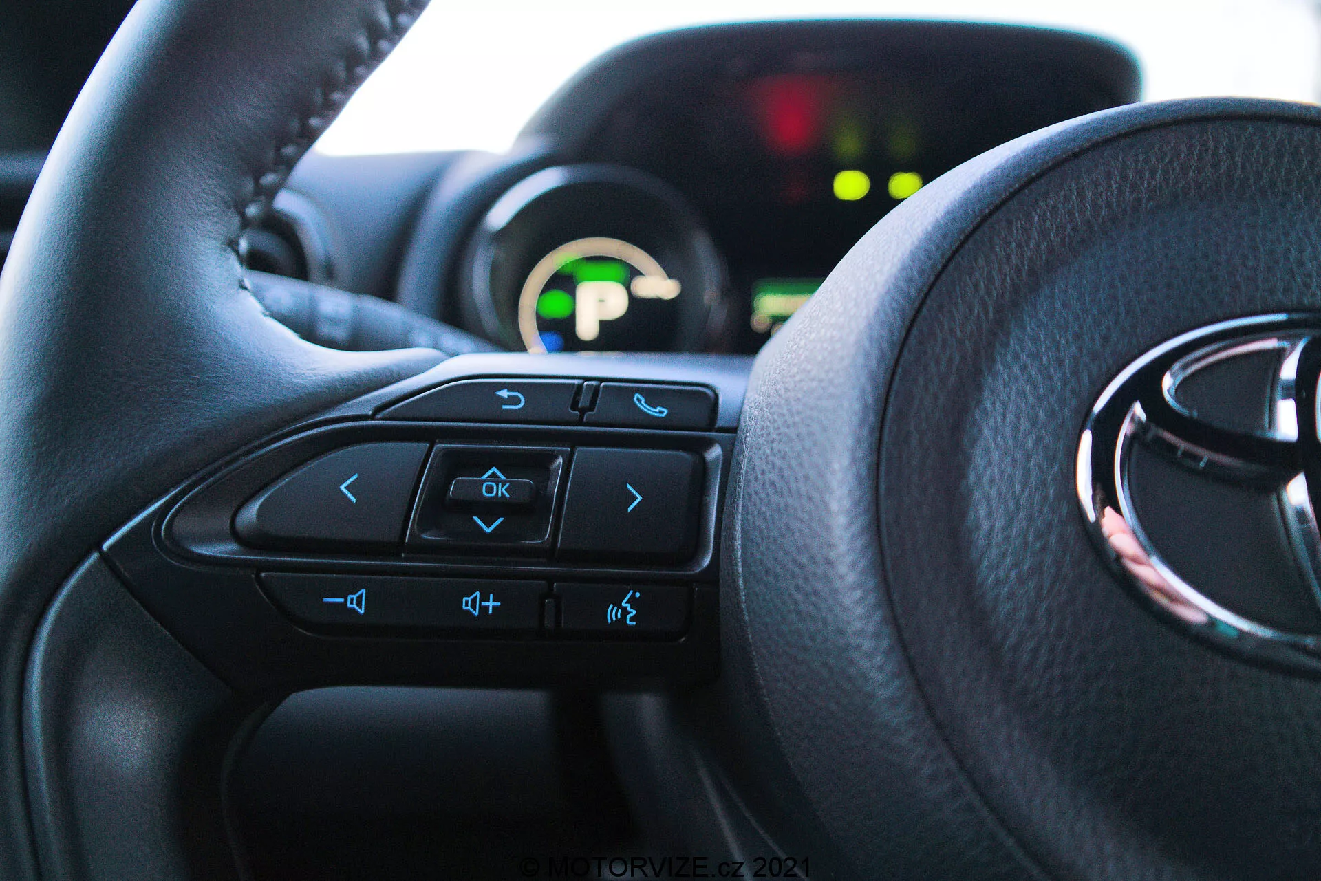 Zbliżony widok wnętrza Toyota Yaris 2020 z perspektywy siedzenia kierowcy, podkreślający kierownicę z wielofunkcyjnymi przyciskami i widocznym w tle zestawem wskaźników. Kierownica dominuje dolne dwie trzecie obrazu, z widocznymi przyciskami strzałek, poleceń głosowych, głośności i systemu rozrywki. Częściowo widoczny zestaw wskaźników jest podświetlony zielonym wyświetlaczem cyfrowym i jest rozmyty ze względu na płytką głębię ostrości. Światło naturalne oświetla wnętrze, podkreślając komfort i przyjazny dla użytkownika interfejs.