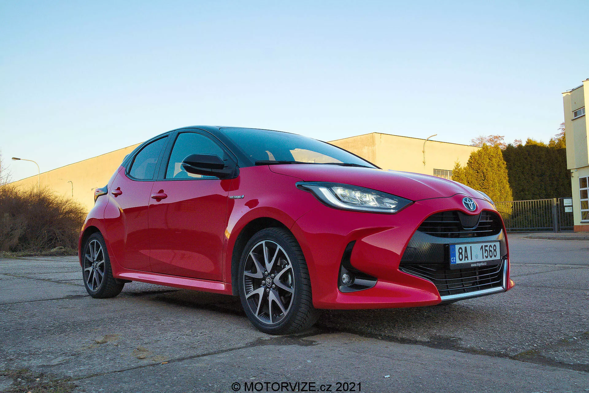 Niskokątny widok trzy-czwarte z przodu wibrującego czerwonego 2020 Toyota Yaris hatchback ukazującego ostre i kątowe reflektory LED lub HID, wyraźną kratkę wlotu powietrza z centralnym emblematem, wieloramienne felgi aluminiowe, zintegrowane kierunkowskazy w lusterkach bocznych, czyste powierzchnie odblaskowe i możliwy system hamulcowy o wysokiej wydajności. Zdjęcie wykonane na ciepło oświetlonym terenie parkingu przemysłowego z znakiem wodnym motorvize.cz 2021.