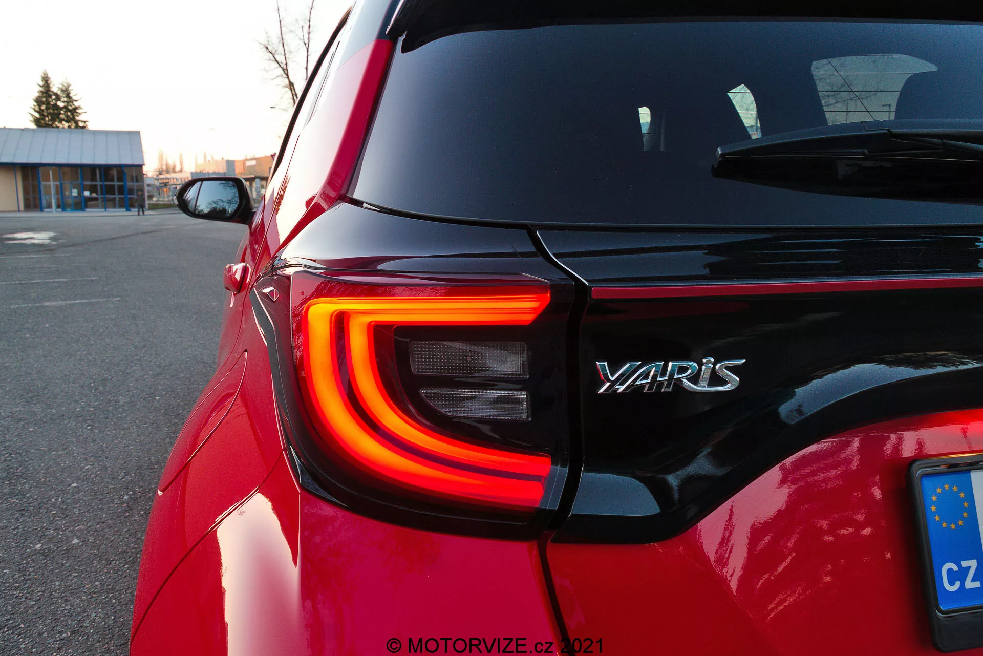 Zbliżenie na tylną część czerwonej Toyota Yaris 2020, ukazujące lewe tylne światło LED z charakterystycznym kształtem litery C, nazwą modelu 'YARIS' na tylnej klapie i tablicę rejestracyjną UE z Czech. Zdjęcie pod kątem ukośnym podkreśla kontury i odblaskowość lakieru samochodu podczas zmierzchu na parkingu, z rozmytym tłem, które skupia uwagę na szczegółach projektu.