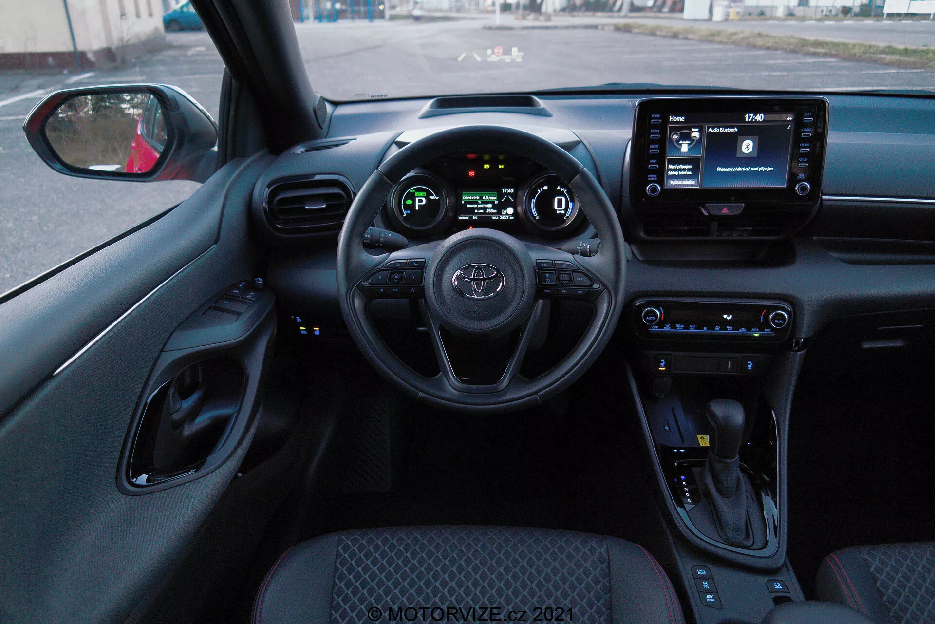 Widok wnętrza z perspektywy fotela kierowcy Toyoty Yaris 2020, z widoczną kierownicą z zamontowanymi przyciskami, cyfrowym panelem zegarów i ekranem multimedialnym. Wnętrze zawiera przyciski sterowania klimatyzacją, dźwignię zmiany biegów oraz przeważnie czarną kolorystykę z teksturowanymi powierzchniami i chromowanymi akcentami.