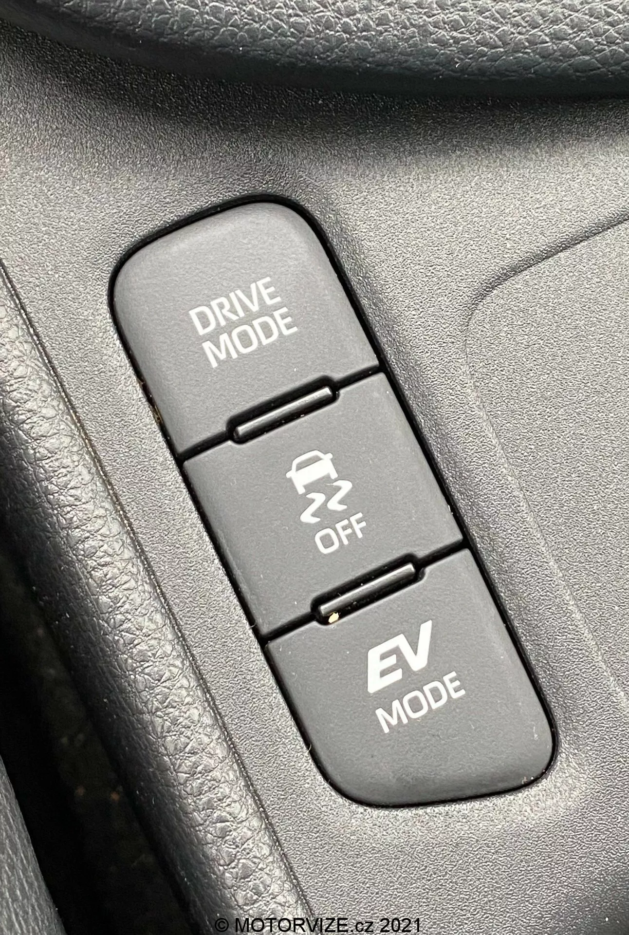 Zbliżenie wnętrza Toyoty Yaris 2020, skupiające się na trzech pionowo ustawionych przyciskach sterujących związanych z trybami jazdy. Od góry do dołu: 'DRIVE MODE' z ikoną krętej drogi, 'Traction Control OFF' z ikoną samochodu w poślizgu oraz 'EV MODE' wskazujący tryb pojazdu elektrycznego. Przyciski są zamontowane na teksturowanej powierzchni, prawdopodobnie części panelu drzwi lub konsoli środkowej. Obraz jest ostro wyostrzony na przyciski, a otaczające tekstury są lekko rozmyte, oświetlone równomiernym światłem dziennym dla jasnej widoczności.
