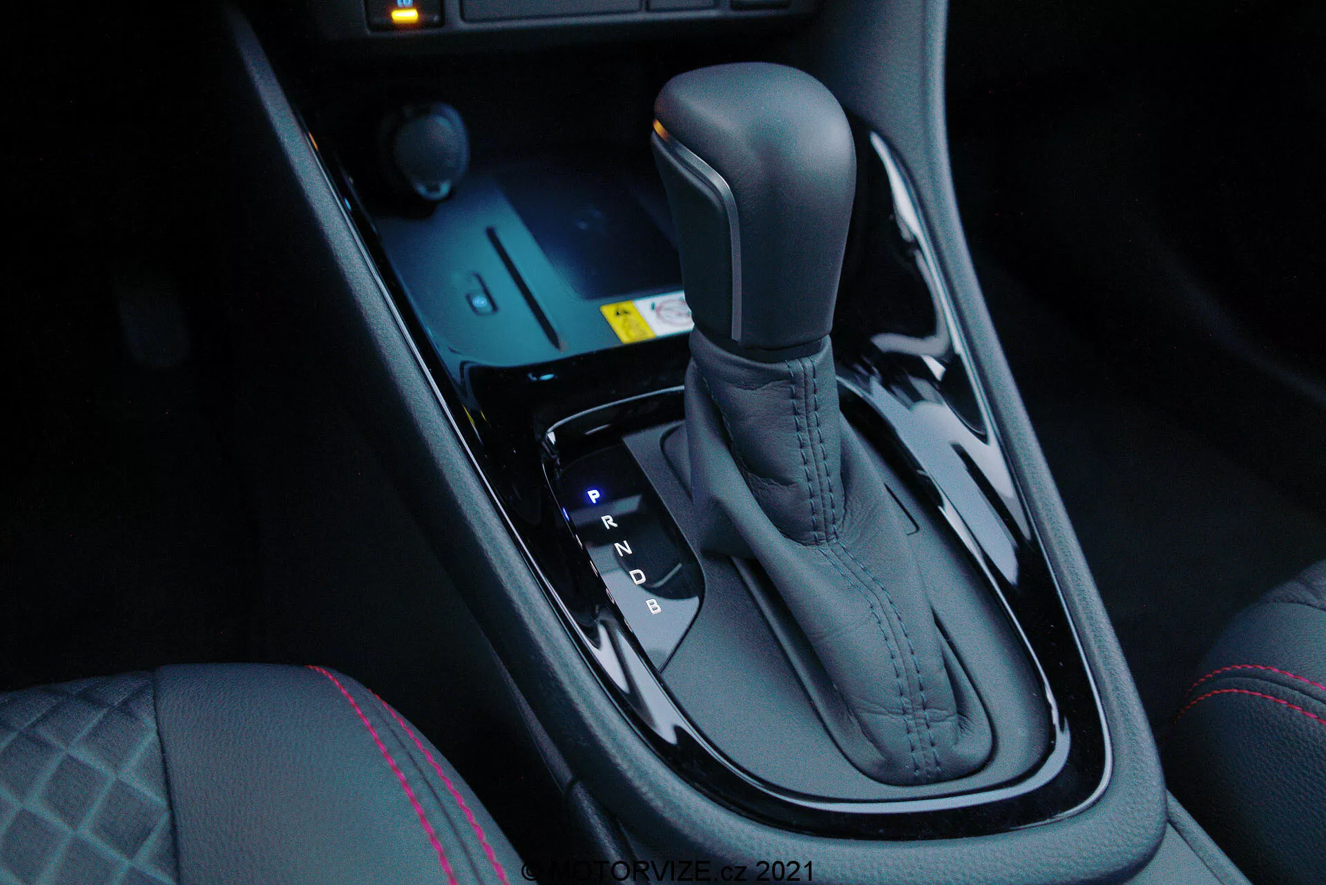 Zbliżenie na automatyczną skrzynię biegów Toyoty Yaris 2020, widziane od strony przedniego pasażera pod kątem w dół, podkreślające eleganckie metalowe wykończenie i ergonomiczny design. Dźwignia zmiany biegów jest umieszczona centralnie między siedzeniami kierowcy i pasażera przedniego i obejmuje pozycje Park, Reverse, Neutral i Drive, a także opcje ręcznej zmiany (+ i -). Przed dźwignią zmiany biegów znajdują się elektroniczne przyciski do trybów jazdy i kontroli trakcji, wraz z podświetlanym na niebiesko przyciskiem startu. Ciemna, błyszcząca konsola centralna dodaje nowoczesnego luksusu, uzupełnionego przez przyciski ogrzewania siedzeń i charakterystyczne szwy na siedzeniu kierowcy, co wskazuje na większy komfort i zaawansowane dynamiki jazdy.