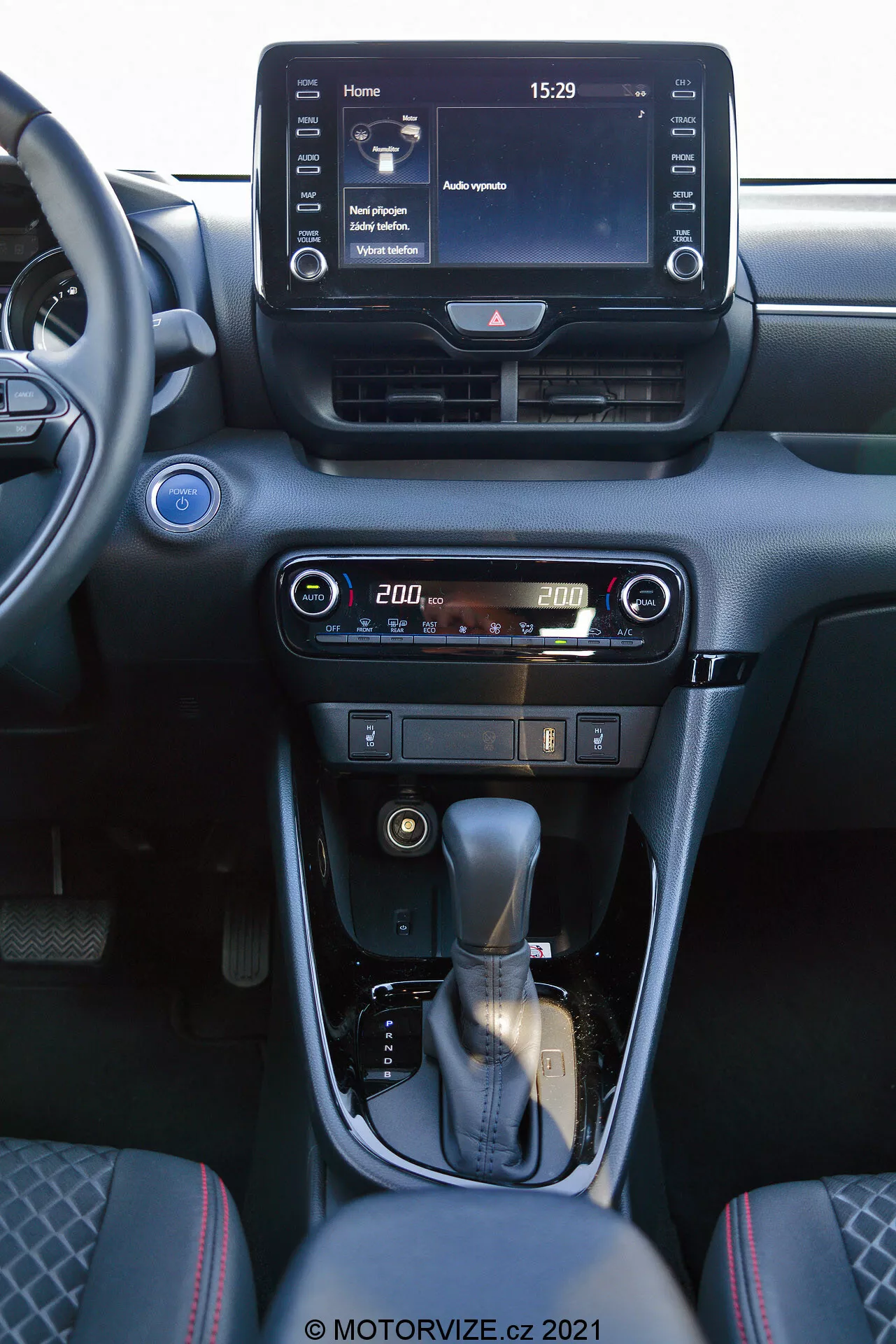 Widok wnętrza Toyota Yaris 2020 skupiający się na konsoli centralnej i elementach deski rozdzielczej. Zdjęcie, wykonane z przedniego siedzenia pasażera, pokazuje cyfrowy system informacyjno-rozrywkowy z ekranem dotykowym wyświetlający ekran główny z czasem 15:29 i różnymi opcjami menu, centralne kratki wentylacyjne oraz przyjazne dla użytkownika sterowanie klimatyzacją i ogrzewaniem z cyfrowym wyświetlaczem temperatury ustawionej na 20,0 stopni. Konsola centralna wyposażona jest w skórzaną dźwignię zmiany biegów z srebrnym akcentem oraz ręczny hamulec. Wnętrze zaprojektowano w ciemnych tonacjach z metalicznymi wykończeniami, tworząc nowoczesną i wyrafinowaną estetykę.