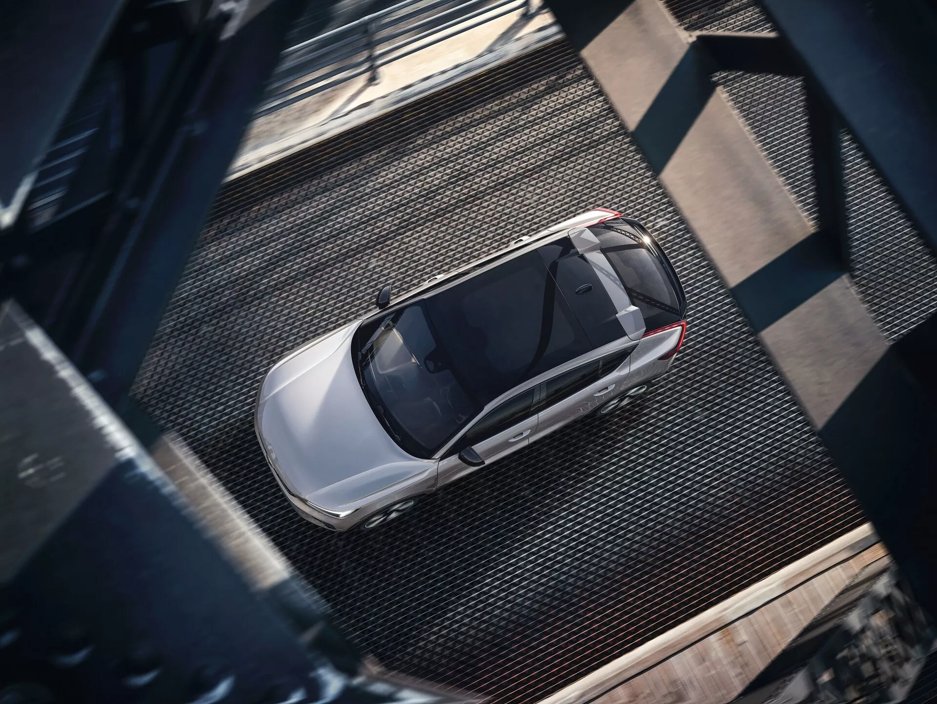 Widok z lotu ptaka na Volvo C40 2021 pierwszej generacji, uchwycony z wysokiej perspektywy. Zdjęcie ukazuje kompletny widok z góry na nowoczesny sedan o eleganckim designie, z metalicznym, błyszczącym wykończeniem w jasnym kolorze, prawdopodobnie srebrnym lub jasno-szarym. Widoczne cechy obejmują dach, maskę, bagażnik, okna oraz fragmenty drzwi, z wyraźnym szyberdachem wskazującym zaawansowaną wentylację. Samochód stoi na teksturowanej, przemysłowej powierzchni z metalowymi siatkami i ciemnymi nitowanymi belkami, rzucając cienie sugerujące boczne oświetlenie, prawdopodobnie rano lub późnym popołudniem.