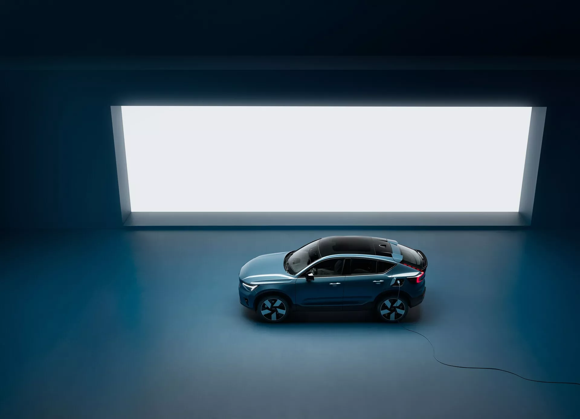 Zdjęcie z wysokiego kąta przedstawiające sedana Volvo C40 2021 z czterema drzwiami i panoramicznym dachem, widziane z góry na ciemnej podłodze z jasnym prostokątnym źródłem światła w tle, prezentujące czysty i minimalistyczny design z ciemnym, błyszczącym nadwoziem i dużymi wieloszprychowymi felgami, podkreślając jego luksusowy i nowoczesny wygląd.