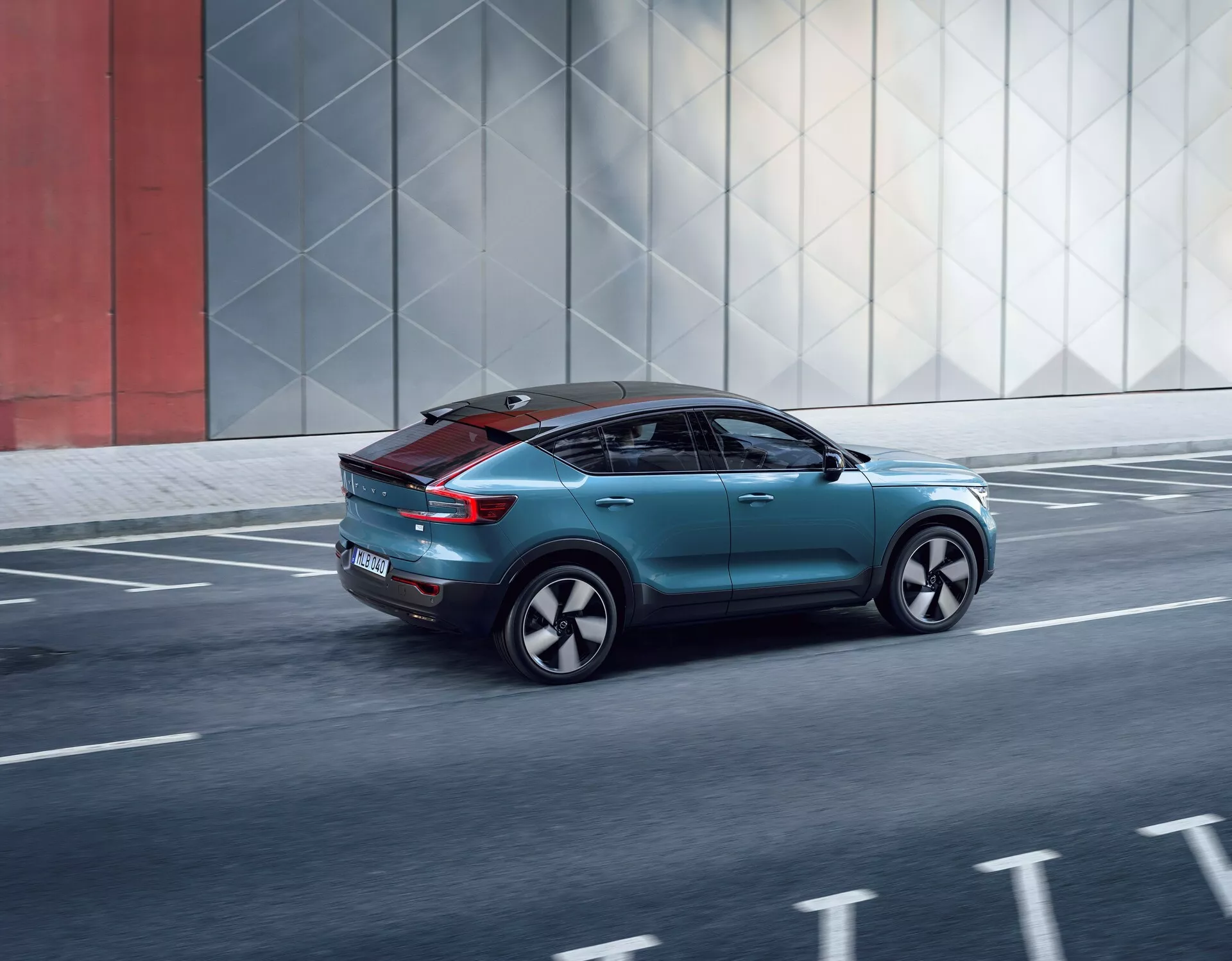 Dynamiczny widok z tyłu od trzech czwartych Volvo C40 2021, ukazujący jego elegancki design, elementy aerodynamiczne, nowoczesne tylne światła, spersonalizowane koła oraz panoramiczny dach, poruszające się wzdłuż miejskiej drogi.