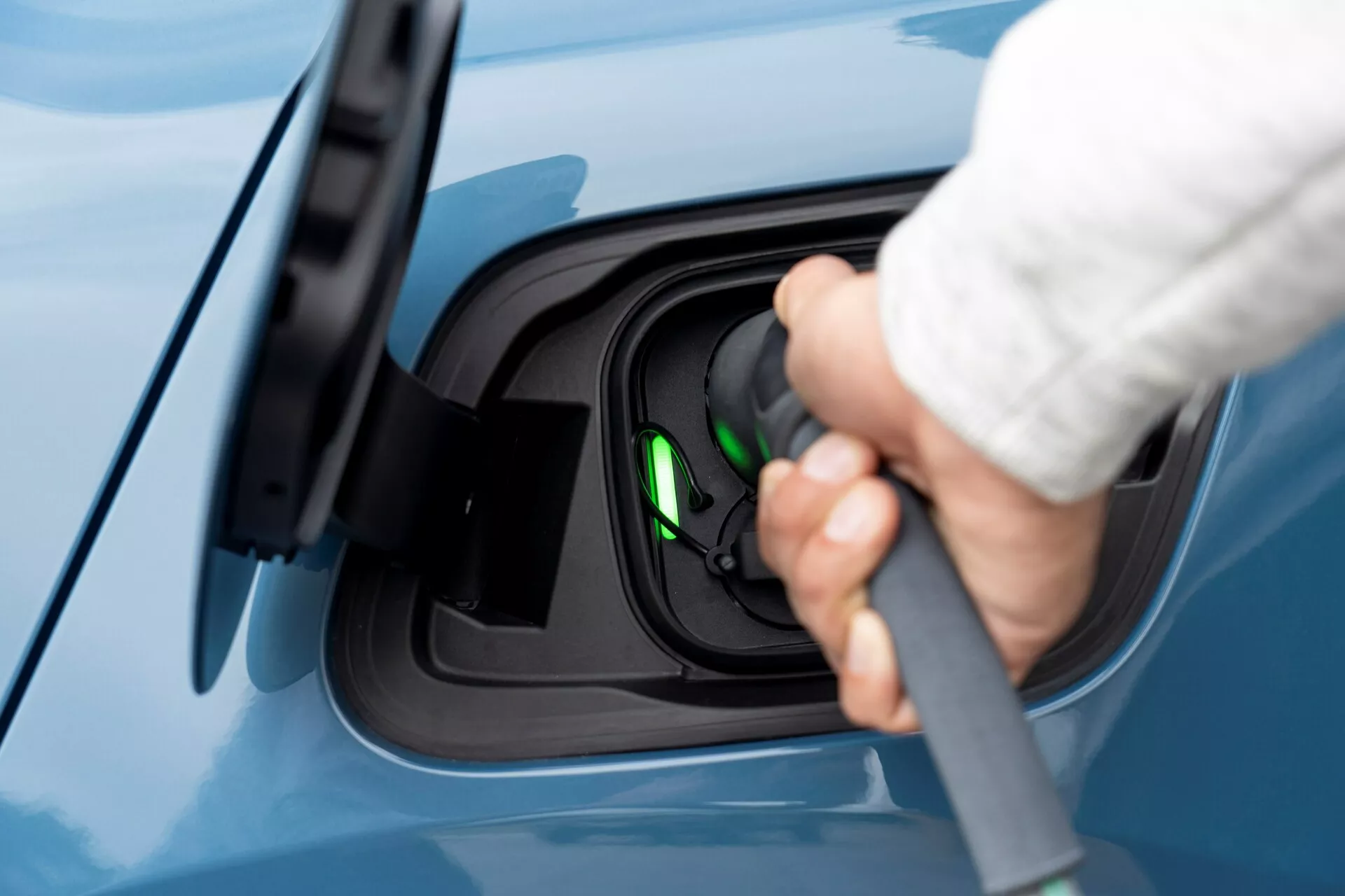 Zbliżony widok z boku na obszar portu ładowania elektrycznego pojazdu Volvo C40 2021 (pierwsza generacja), z ludzką ręką podłączającą kabel ładujący do portu. Obraz koncentruje się na procesie ładowania, podkreślając otwarty port ładowania z prawdopodobnie używanym złączem typu 2. Zielona lampka wewnątrz gniazda wskazuje, że pojazd jest gotowy do ładowania lub jest aktualnie ładowany. Widok jest lekko podwyższony, pokazując nieskazitelne niebieskie wykończenie lakieru na samochodzie, podkreślając czysty i nowoczesny wygląd pojazdu elektrycznego.