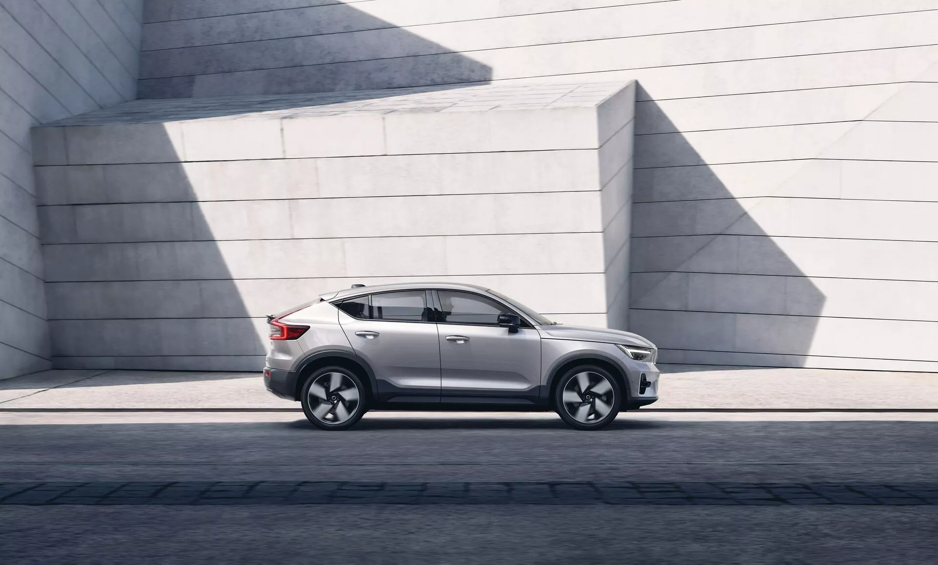 Obraz nowoczesnego SUV Volvo C40 2021 zaparkowanego równolegle, pokazujący lewy profil boczny od lewego przedniego reflektora do lewego tylnego światła. Kąt podkreśla prześwit i postawę na utwardzonej powierzchni. Światło słoneczne tworzy cienie na błyszczącej powłoce lakierniczej. SUV ma elegancki design z delikatnie opadającą linią dachu, wyraźnymi konturami drzwi, przyciemnianymi szybami i współczesnymi lusterkami bocznymi. Aluminiowe koła z wieloramiennym wzorem i opony o niskim profilu wskazują na osiągi i luksus. Widoczne są listwy progowe, formy nadkoli i linia charakterystyczna biegnąca przez klamki drzwi, sugerując solidność. Brak widocznego wydechu sugeruje możliwy napęd elektryczny lub hybrydowy. Minimalistyczne betonowe struktury w tle kontrastują z wyszukanymi krzywiznami samochodu, podkreślając jego miejski i designerski wygląd.