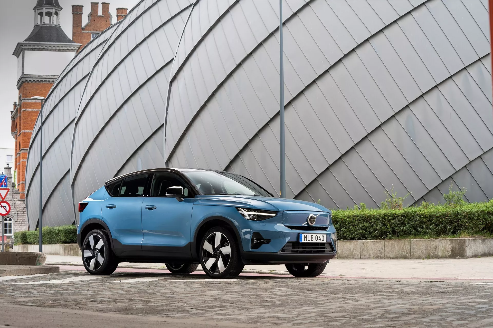 Nowoczesne niebieskie SUV Volvo C40 2021 zaparkowane na utwardzonym obszarze miejskim z kanciastą, metaliczną strukturą w tle. Zdjęcie wykonane z niskiego kąta od strony lewego przodu ukazuje przednią kratkę pojazdu, reflektory LED, aerodynamiczny profil, wieloramienne felgi aluminiowe, lusterka boczne z wbudowanymi kierunkowskazami, matowe lub satynowe wykończenie lakieru, przyciemniane szyby, przednią tablicę rejestracyjną i rzeźbione linie boczne nadwozia.
