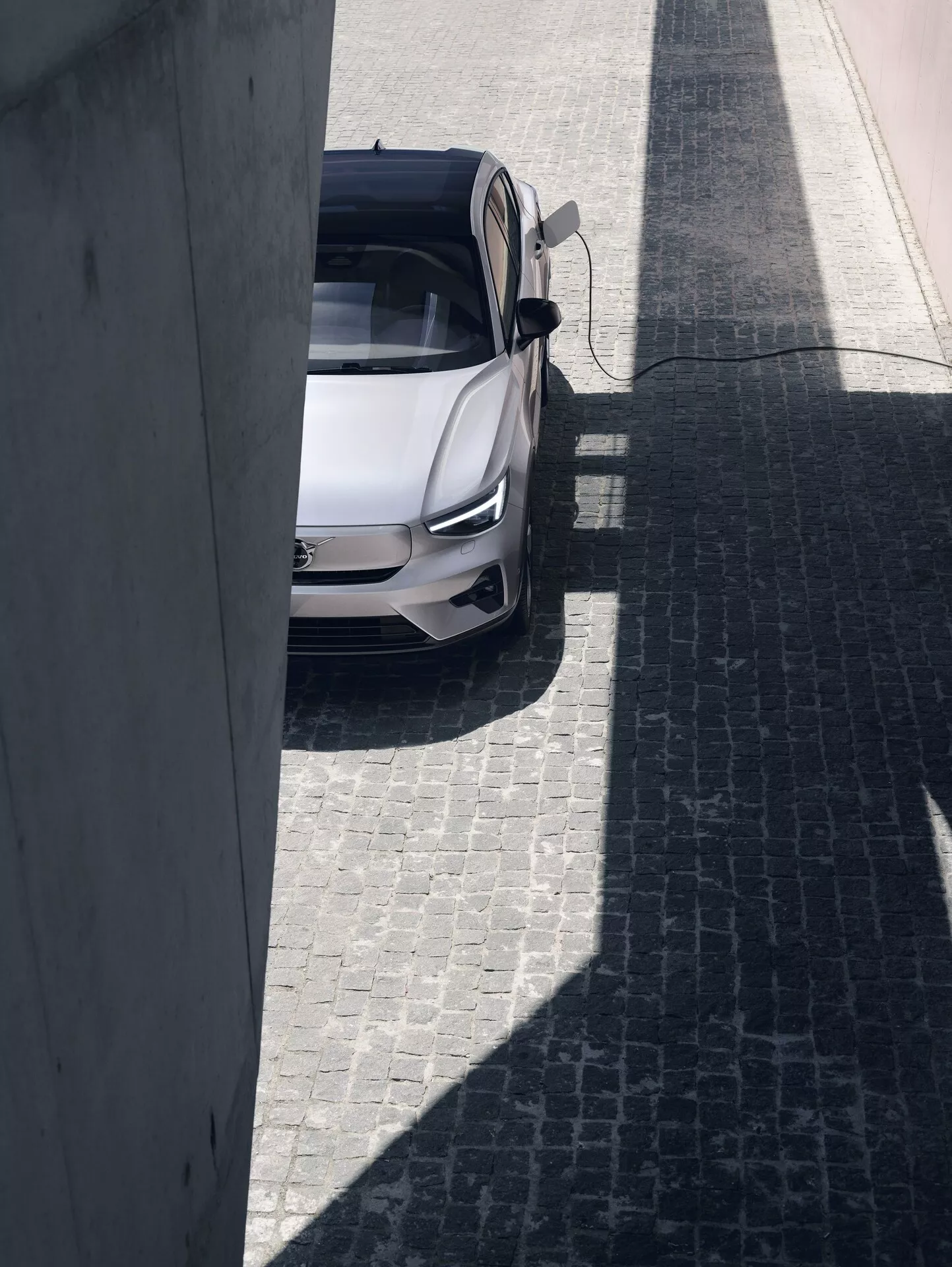 Widok z lotu ptaka na Volvo C40 2021 pierwszej generacji, sfotografowane w słoneczny dzień z podwyższonej perspektywy. Samochód jest wyśrodkowany na brukowanej ulicy, ma jasny kolor, prawdopodobnie biały lub srebrny, oraz panoramiczny dach. Otwarte drzwi po stronie kierowcy umożliwiają wgląd do wnętrza, podkreślając nowoczesny, sportowy design sedana z potencjalnie zaawansowanymi funkcjami automotive.