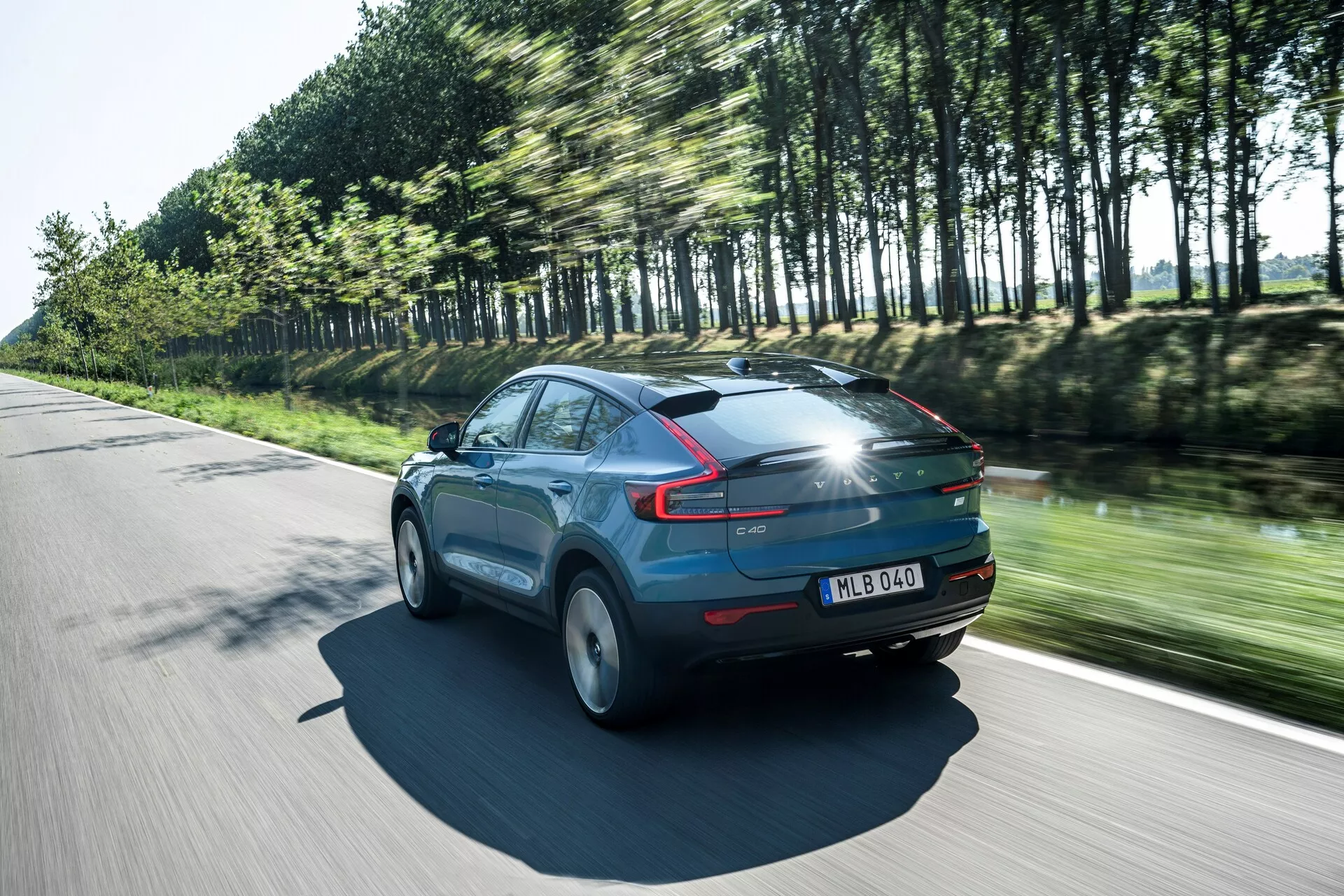 Dynamiczne ujęcie Volvo C40 z 2021 roku (1. generacja) w ruchu w słoneczny dzień, widok trzy czwarte od tyłu, podkreślający profil pojazdu i jego aerodynamikę. Samochód, pomalowany błyszczącym niebieskim kolorem, posiada charakterystyczne tylne światła LED, rozmyte w ruchu koła i drzewa odbijające jego prędkość. Godne uwagi szczegóły to panoramiczny dach, tylny spojler i tablica rejestracyjna na tylnej klapie, wyróżniające nowoczesne elementy designu i zaawansowaną technologię oświetleniową.