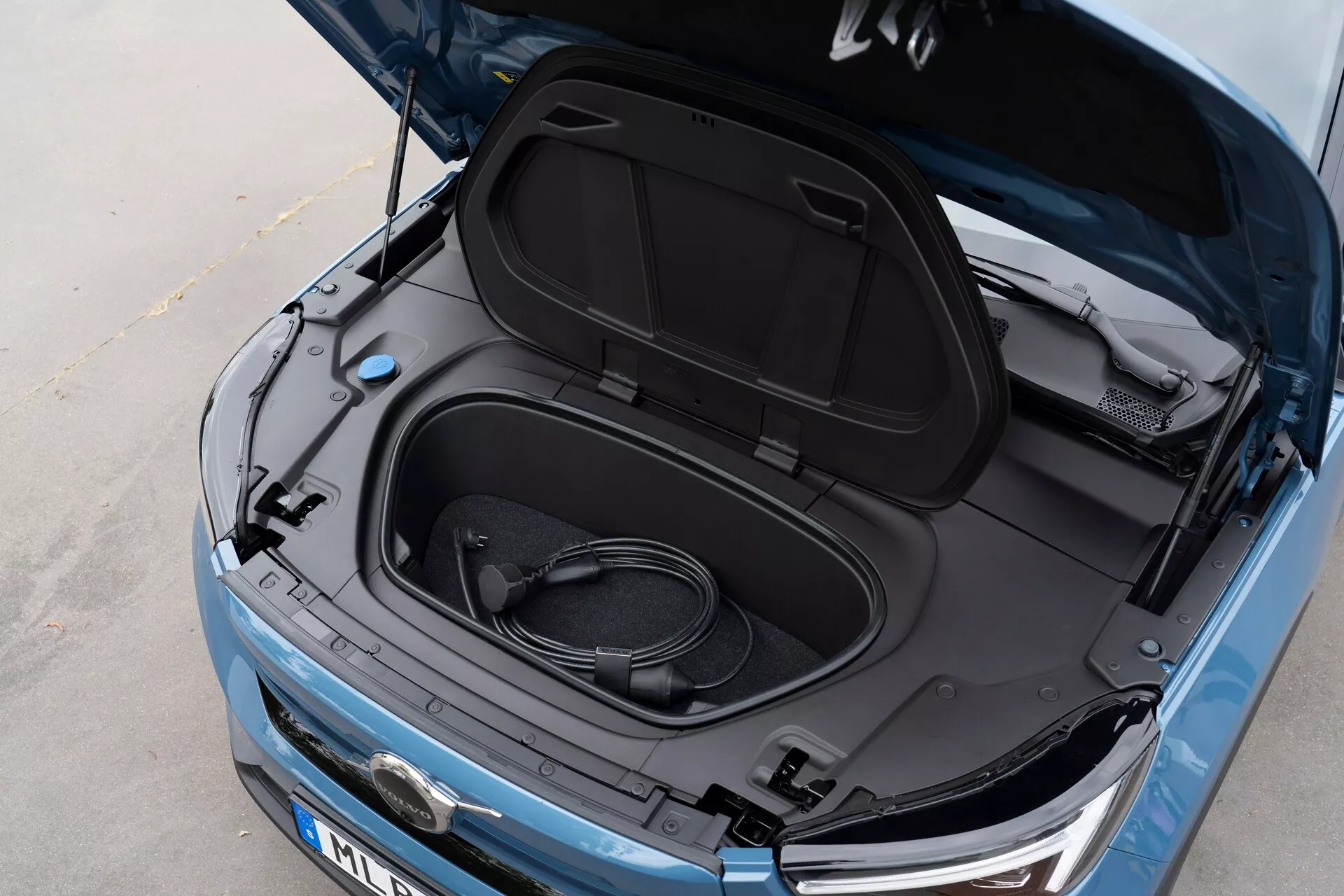 Zbliżenie pod kątem z góry na Volvo C40 2021 (1. generacja) z otwartym frunkiem, ukazującym pustą przestrzeń ładunkową i zwinięty kabel do ładowania, wskazującym, że jest to pojazd elektryczny. Metaliczny niebieski lakier, czarne wnętrze pokrywy frunka z informacyjnymi naklejkami, częściowy widok na logo producenta, eleganckie reflektory i widoczny przedni grill, sugerują, że EV ma nowoczesną technologię LED i opływową estetykę.
