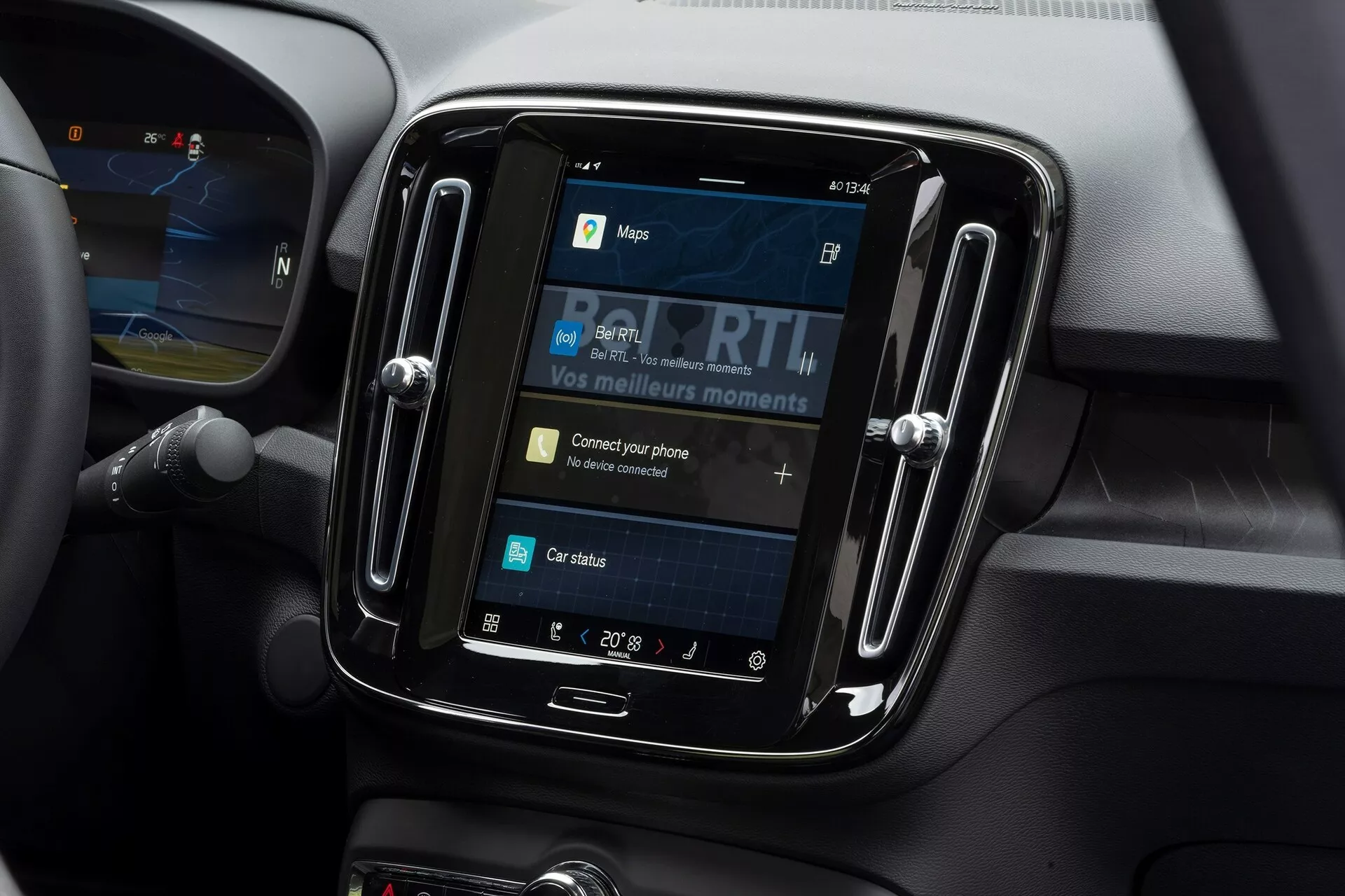 Zbliżenie systemu infotainment wewnątrz Volvo C40 2021. Zdj cie wykonane z niewielkiego kąta w dół skupia się na wyświetlaczu dotykowym umieszczonym w błyszczącym, czarnym obramowaniu. Ekran pokazuje opcje takie jak mapy, media, łączność telefoniczna i status samochodu, z interaktywnym elementem 'Podłącz swój telefon'. Wokół systemu infotainment widać miękkie materiały i linie konturowe wskazujące na kratki wentylacyjne, co sugeruje nowoczesne wnętrze samochodu. Ustawienie temperatury pokazuje 20° Celsjusza, a po lewej stronie widać część kierownicy.