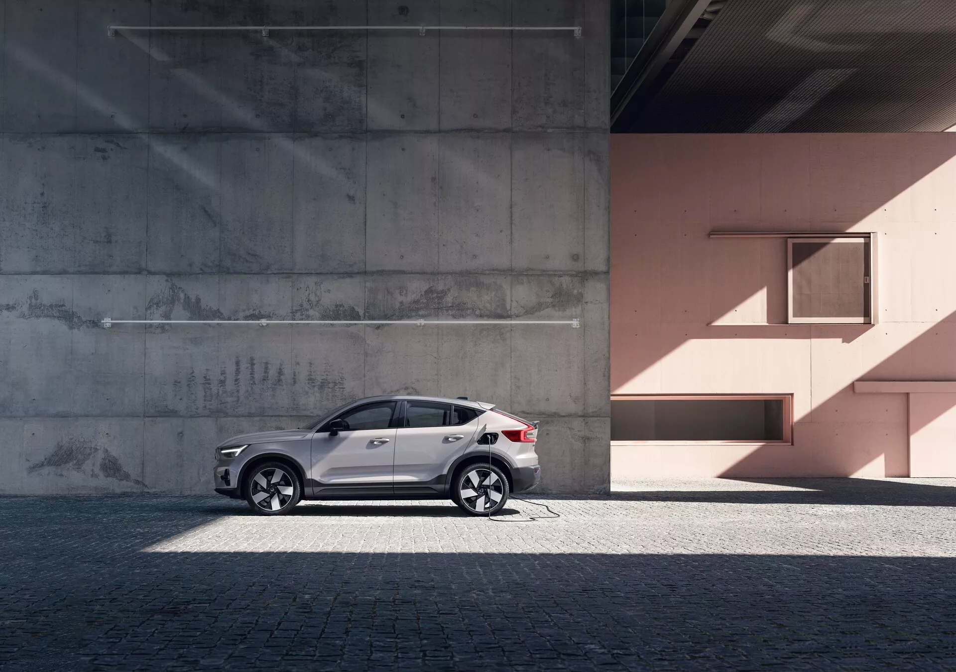 Profil boczny eleganckiego SUV-a Volvo C40 z 2021 roku (1. generacja) na tle miejskim z betonowymi i brzoskwiniowymi ścianami. Samochód ma odblaskowy biały lakier, stylowe aluminiowe felgi, przyciemniane szyby, srebrne relingi dachowe i zintegrowane kierunkowskazy w lusterkach. Zdjęcie podkreśla linie i krzywizny projektu pojazdu w naturalnym świetle dziennym, dodając poczucie luksusu i wyrafinowania.