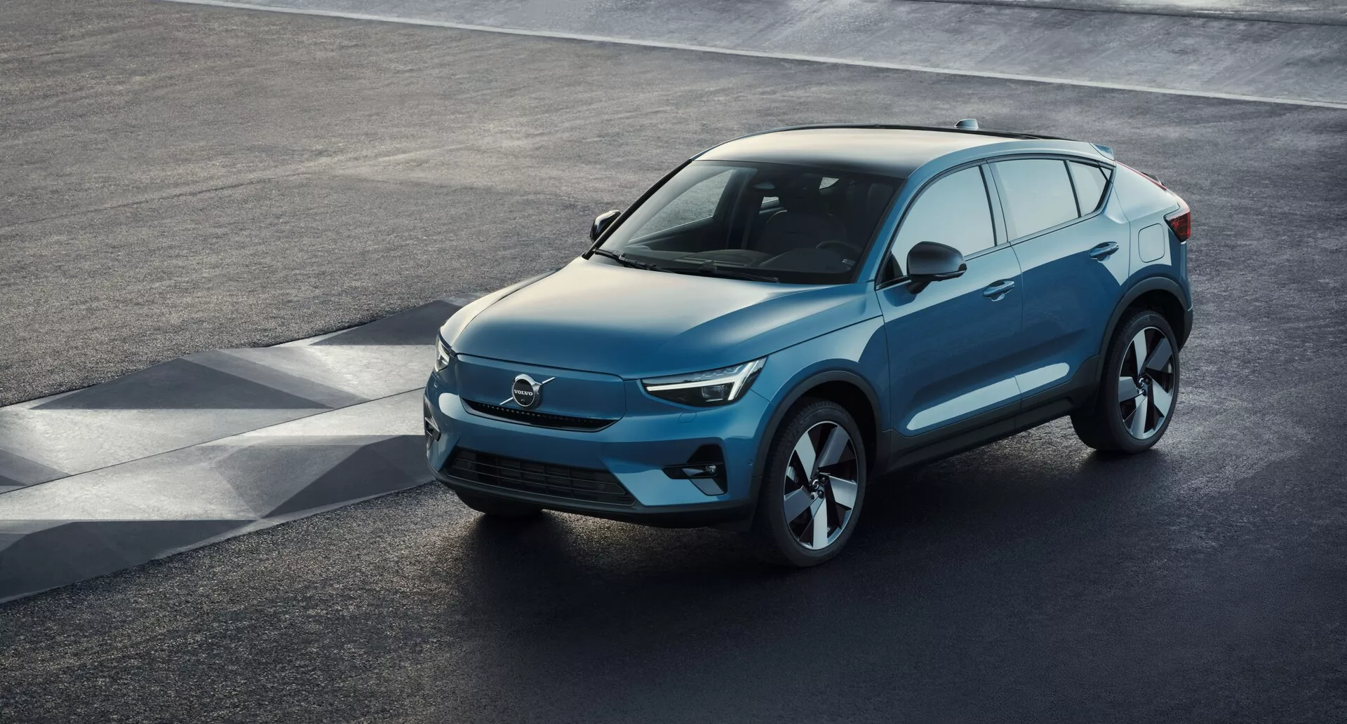 Widok z podwyższenia na nowoczesny SUV Volvo C40 2021 (1. generacja) zaparkowany na asfalcie, ukazujący wysokokątowy, trzyczęściowy widok przedni. Elegancki przód, prawdopodobnie charakterystyczny dla pojazdów elektrycznych z minimalnymi wlotami powietrza, łączy się z ostrymi reflektorami LED, gładko opadającą maską i konturowanym profilem bocznym z dużymi aluminiowymi felgami. Lusterka boczne z wbudowanymi kierunkowskazami, duża powierzchnia okien z możliwym przyciemnieniem oraz łagodnie opadająca linia dachu z relingami dachowymi do użytku to godne uwagi cechy. Wyraźny widok uchwytuje luksusowy design pojazdu, zaawansowaną aerodynamikę i nowoczesne elementy technologiczne.