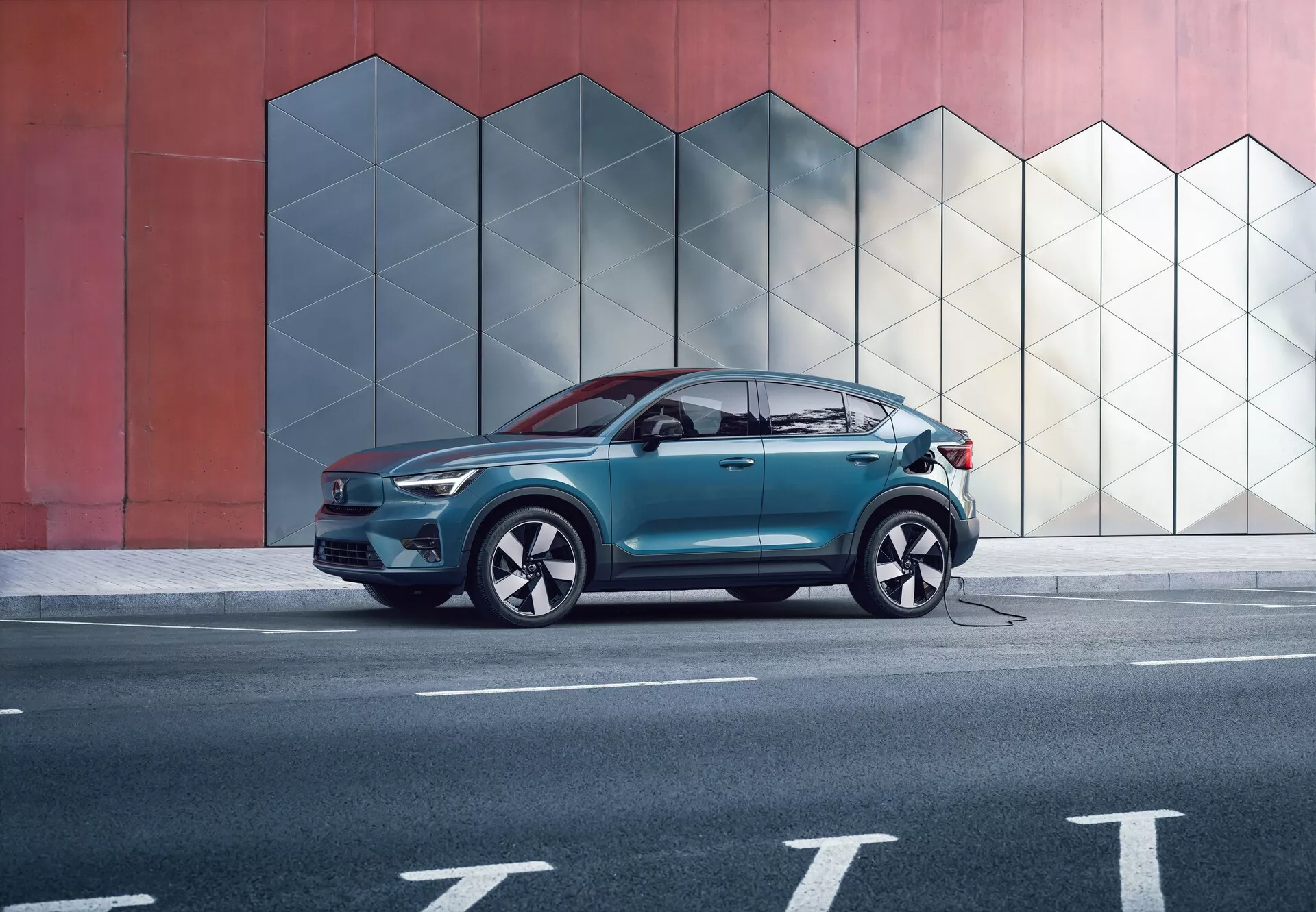Profilowy widok boczny nowoczesnego SUV-a Volvo C40 z 2021 roku (1. generacja) przed miejską ścianą o geometrycznym wzorze, uchwycony z średniej odległości. Obraz podkreśla eleganckie aerodynamiczne linie nadwozia, okna z chromowanymi listwami i duże koła z niskoprofilowymi oponami. SUV ma podłączony kabel ładujący z boku, wskazując na technologię pojazdu elektrycznego. Na zdjęciu nie ma osób, co podkreśla projekt i cechy samochodu.
