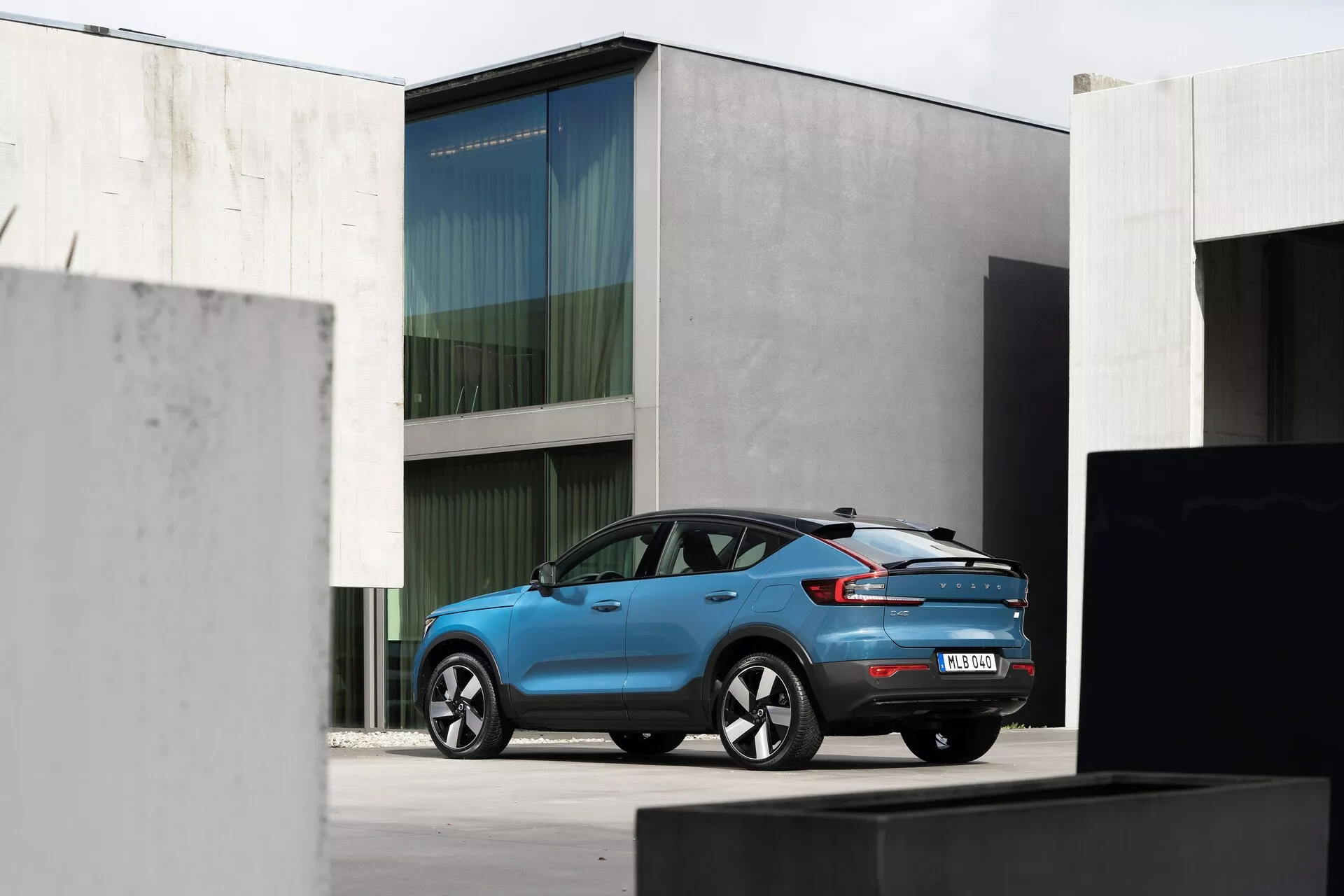 Widok z tyłu pod kątem samochodu Volvo C40 2021 (1. generacja) w jasnoniebieskim kolorze, pokazany w miejskim otoczeniu z minimalistyczną architekturą. Samochód posiada wydłużone tylne światła LED, zintegrowany tylny spojler, antenę w kształcie płetwy rekina, aluminiowe felgi o charakterystycznym wzorze szprych oraz czujniki na tylnym zderzaku. Naturalne, rozproszone oświetlenie podkreśla nowoczesny design.