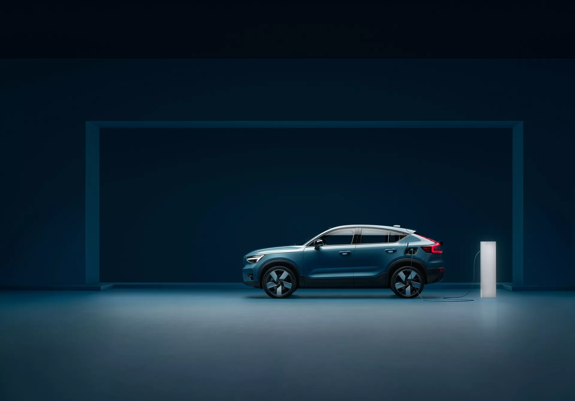 Widok boczny nowoczesnego samochodu Volvo C40 2021 (1. generacja) w studio z refleksyjną podłogą, pokazujący elegancki design, ciemnoniebieską metaliczną farbę, opadającą linię dachu, duże wieloramienne felgi aluminiowe, zarysowane linie, solidną konstrukcję, zintegrowane lusterka boczne z kierunkowskazami, tylne światła LED i port ładowania elektrycznego.
