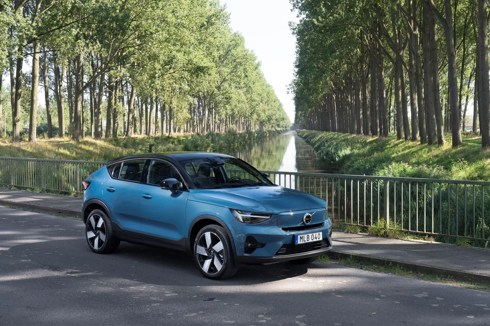 Volvo C40 2021 (Pierwsza Generacja) kompaktowy SUV w widoku trzy-czwarte od przodu-w lewo na asfaltowej drodze. Prezentuje nowoczesne elementy designu, takie jak wyrazisty grill z centralnym emblematem, eleganckie światła LED, stylowe felgi aluminiowe, lusterka boczne z wbudowanymi kierunkowskazami, aerodynamicznie ukształtowane nadwozie z jaskrawo niebieskim lakierem, przyciemniane szyby i cztery drzwi. Ustawiony w zewnętrznym środowisku z drzewami wzdłuż drogi, skoncentrowany na estetyce i cechach technologicznych pojazdu.